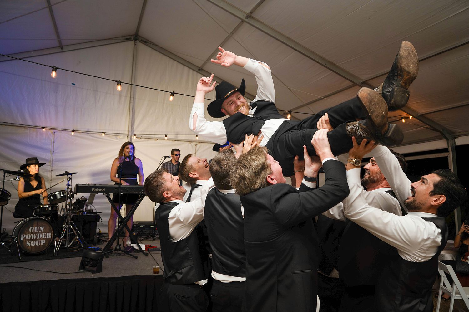 Groomsmen lifting groom in the air at a wedding reception with a live band performing on stage.