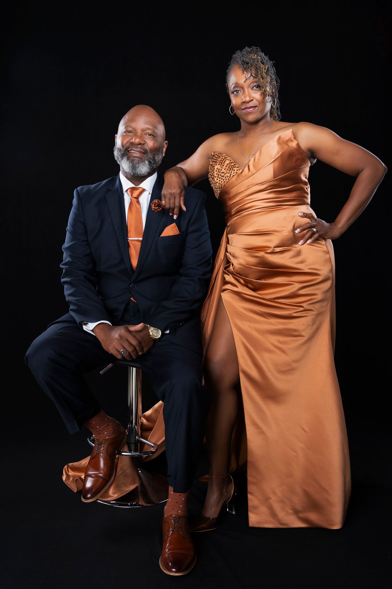 Glamorous couple poses together in coordinated formal wear featuring orange tones against dark backdrop.