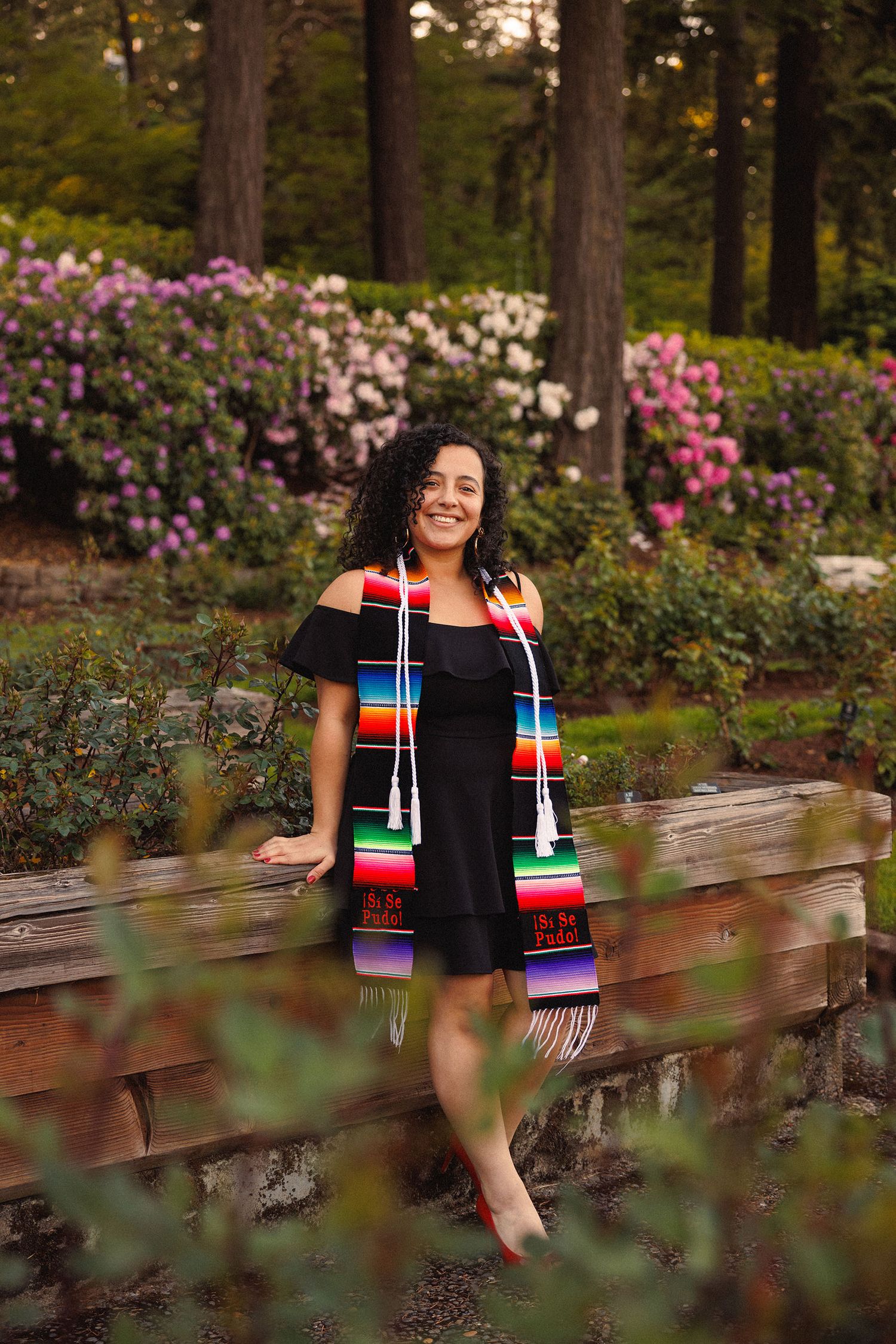 Graduate in a colorful striped graduation stole stands smiling near blooming rhododendrons in a garden setting.
