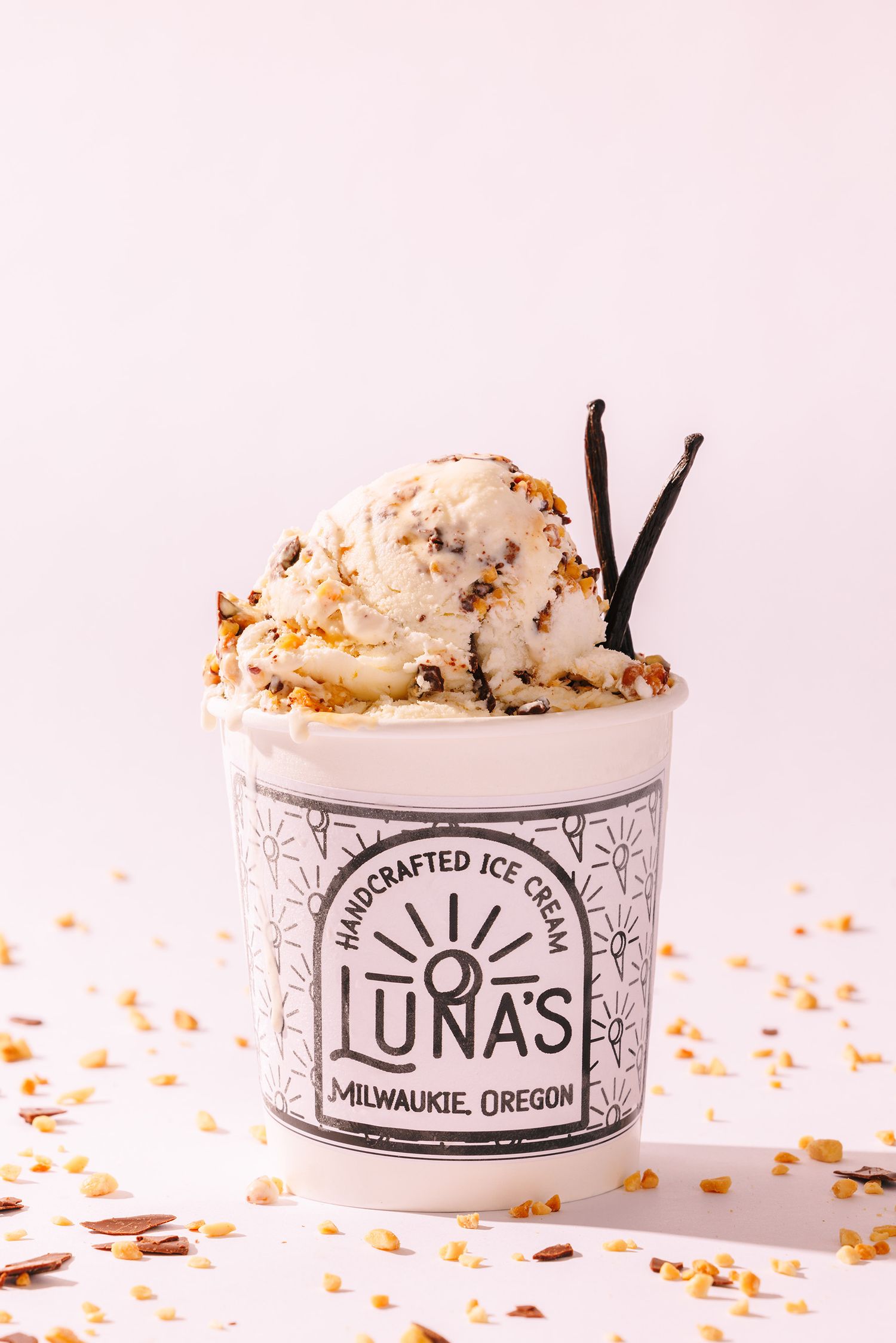 A scoop of vanilla ice cream with toppings in a Luna's branded paper cup, scattered with crumbs against a pink background.