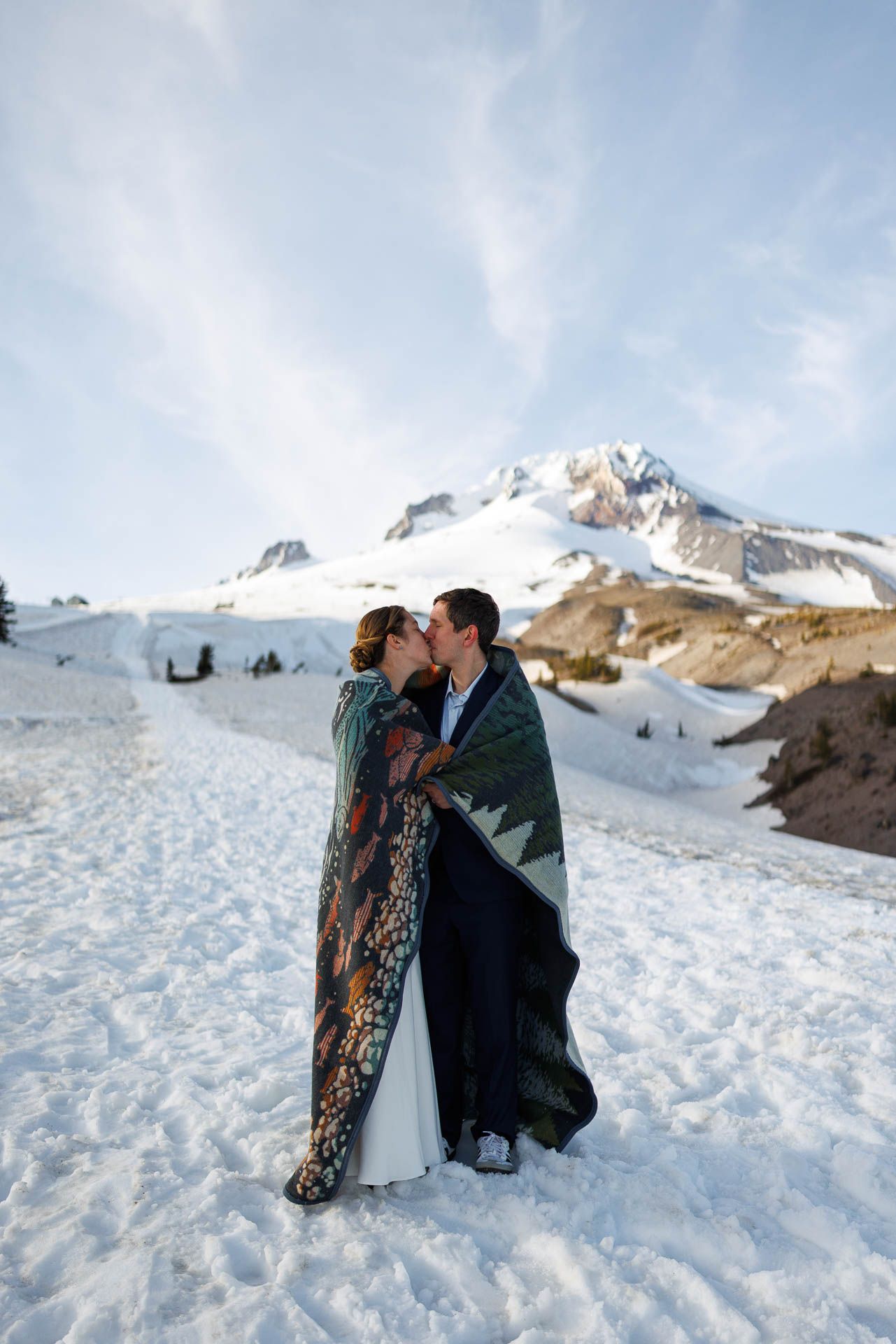 A couple wrapped in a blanket share an intimate moment in the snow with Mount Hood's snowy peak in the background.