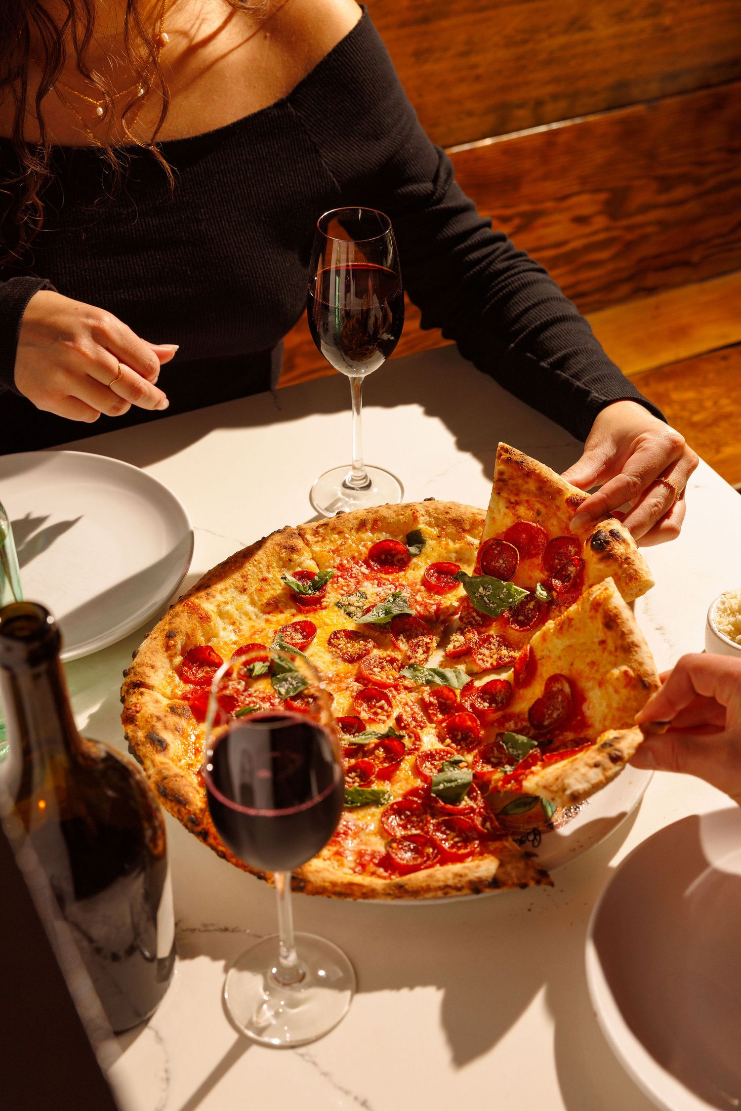 Overhead view of pizza at a restaurant table with red wine glasses and someone reaching for a slice on wooden surface.