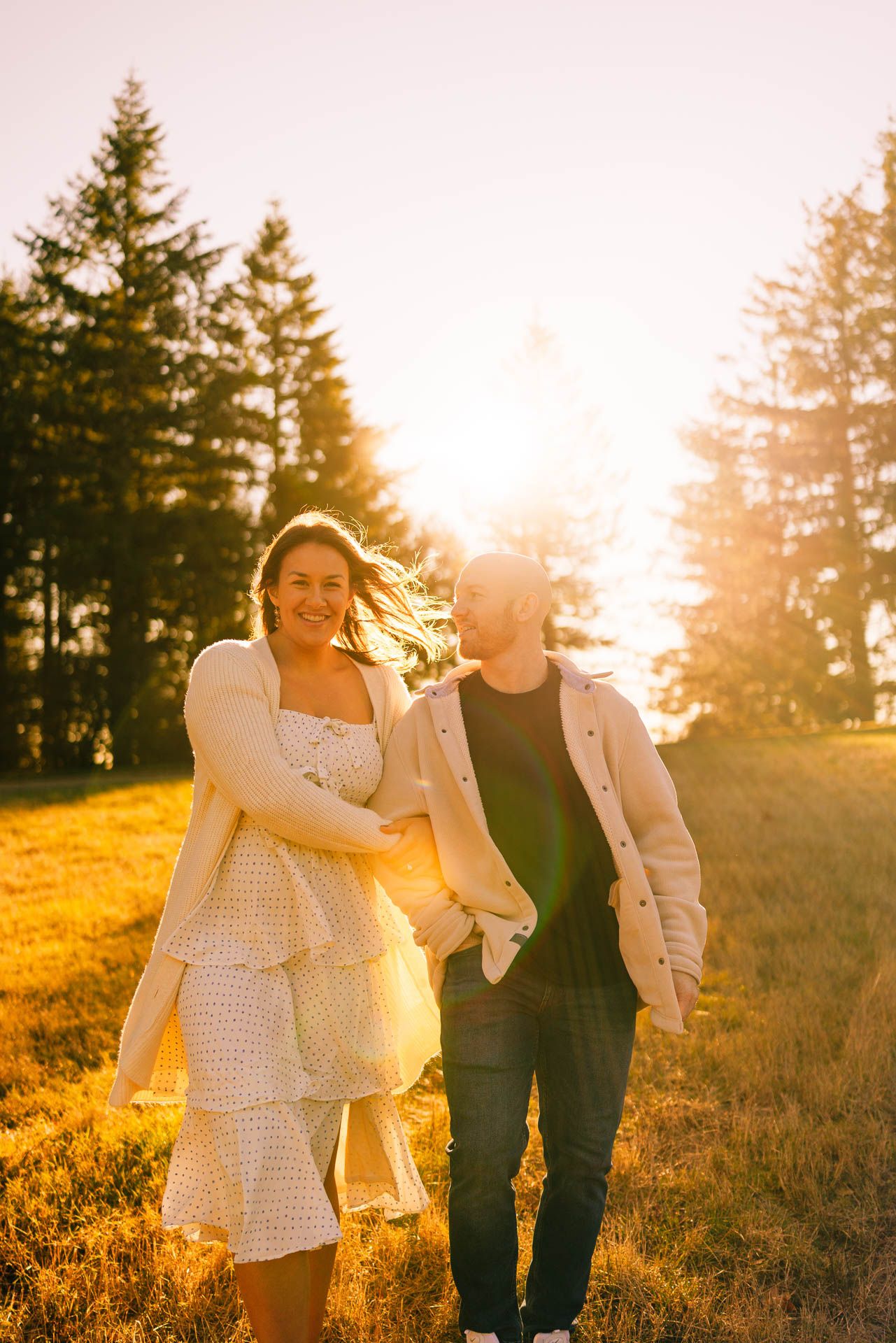 Two people walking through a golden field at sunset as sunlight filters through tall pine trees creating a warm atmosphere.