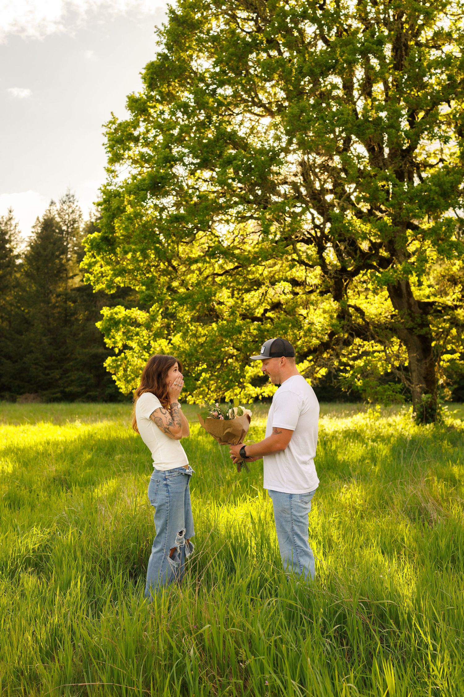 Two people stand together in a sunlit meadow with tall grass and a majestic oak tree during golden hour.