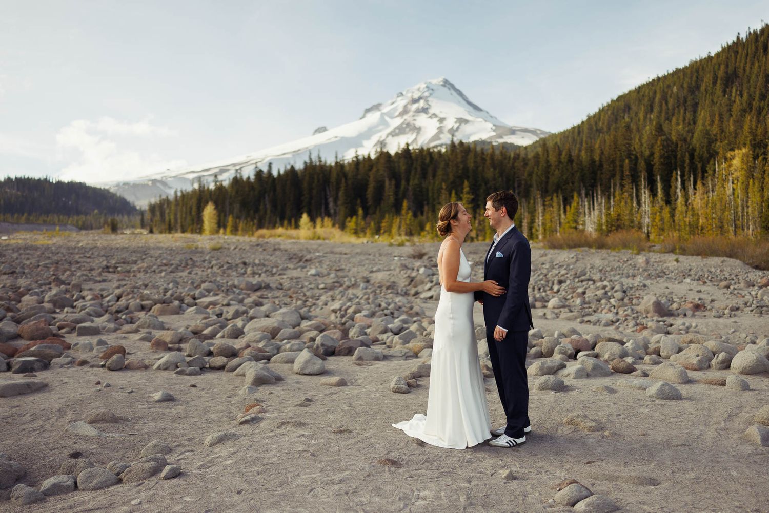 A couple stands on a rocky riverbed with Mount Hood and evergreen forest in the background in Oregon.