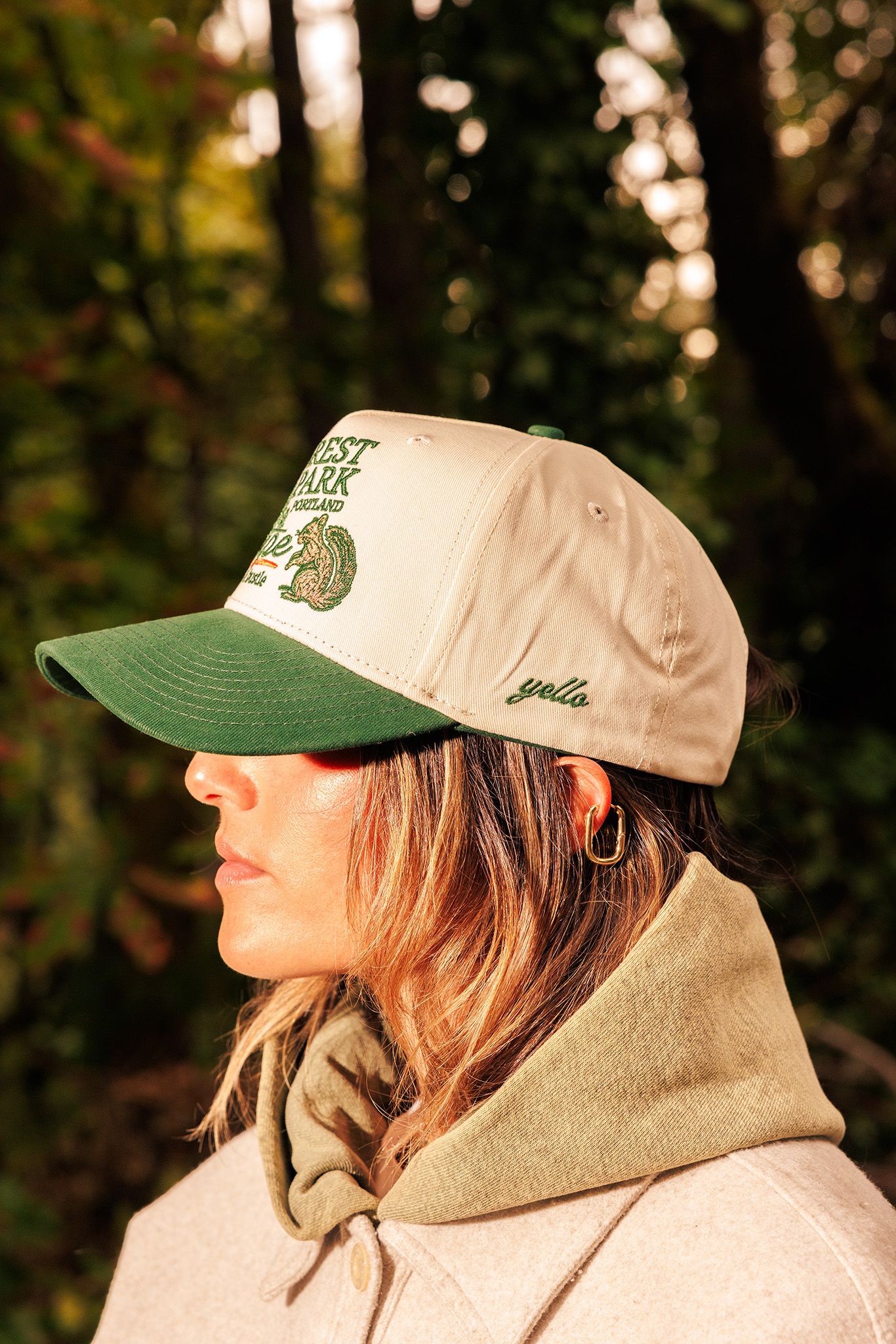 A side profile view showing a person wearing a beige hoodie and baseball cap with green accents in a forest setting.