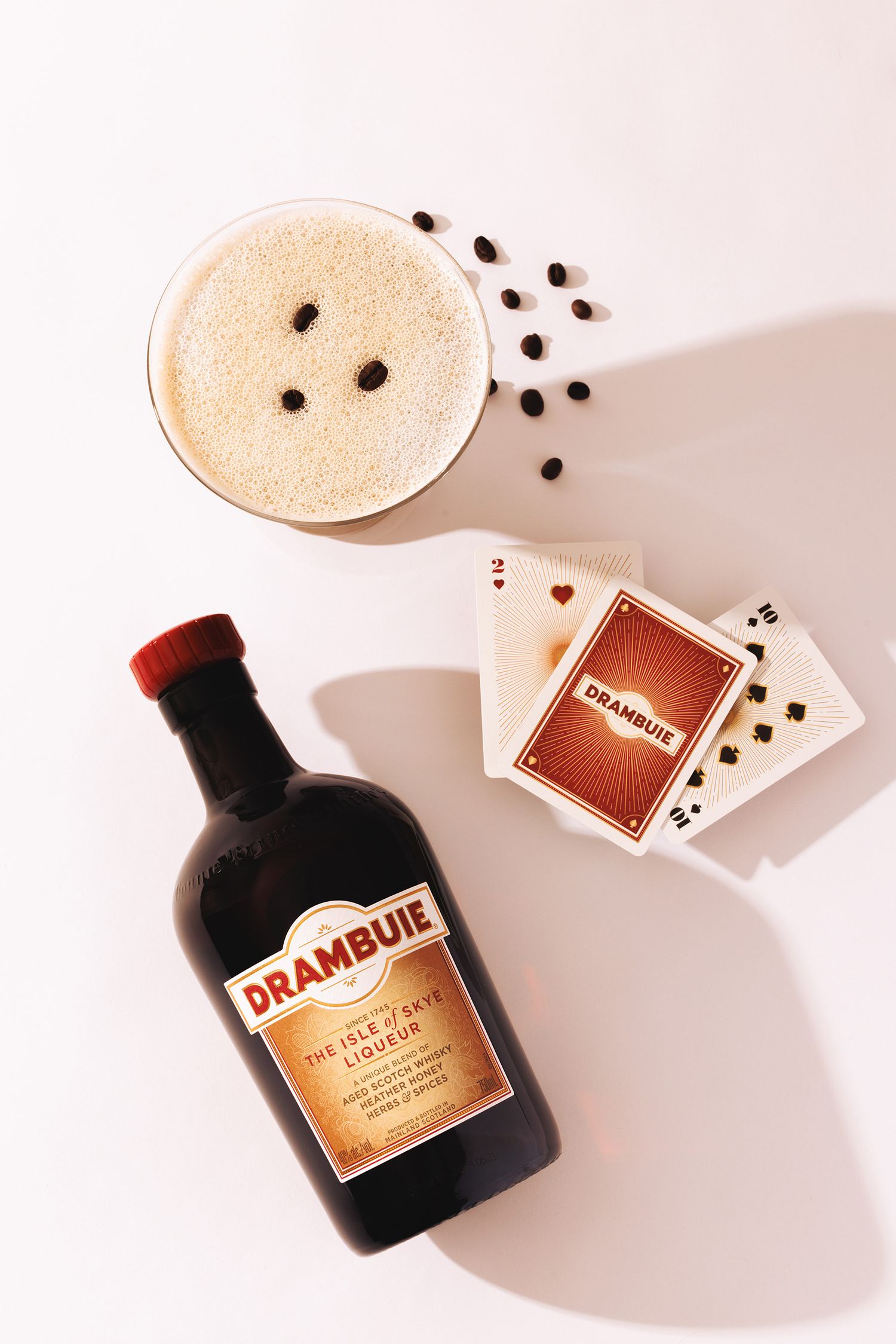 Bottle of Drambuie liqueur with a cocktail glass and playing cards arranged on a light pink surface with coffee beans.