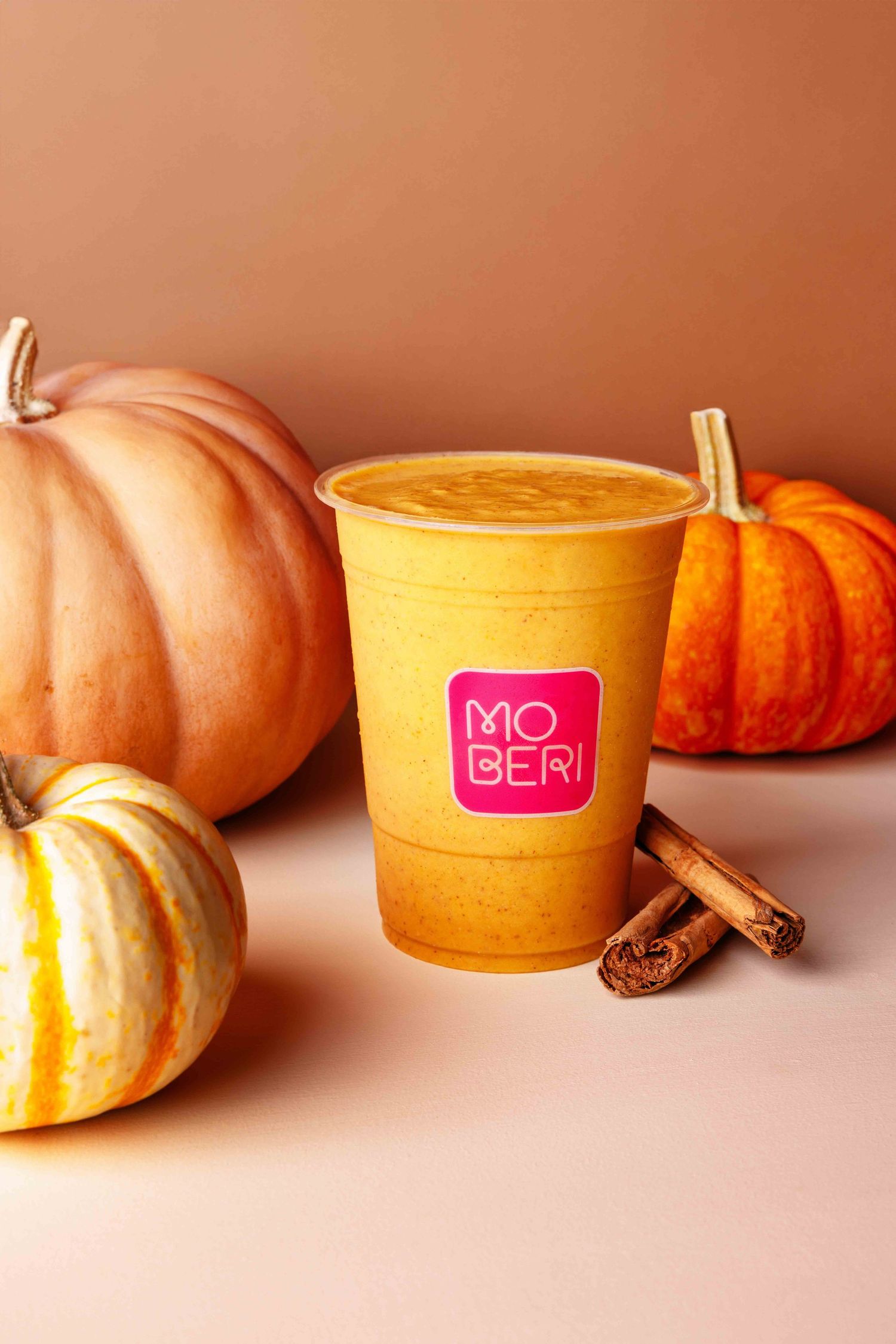 Orange pumpkins and miniature gourds arranged with pink candle holders and cinnamon sticks on a beige surface.