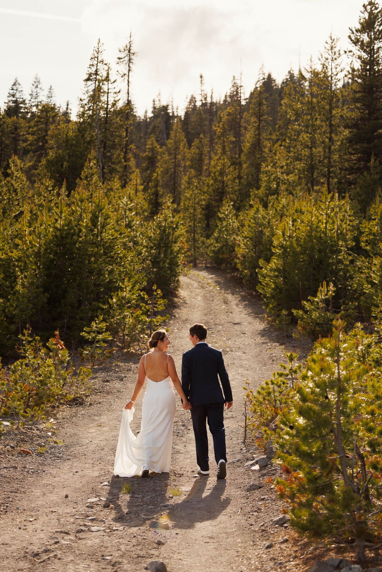 Couple walking down a dirt path surrounded by evergreen trees and golden sunlit foliage in a mountain forest.