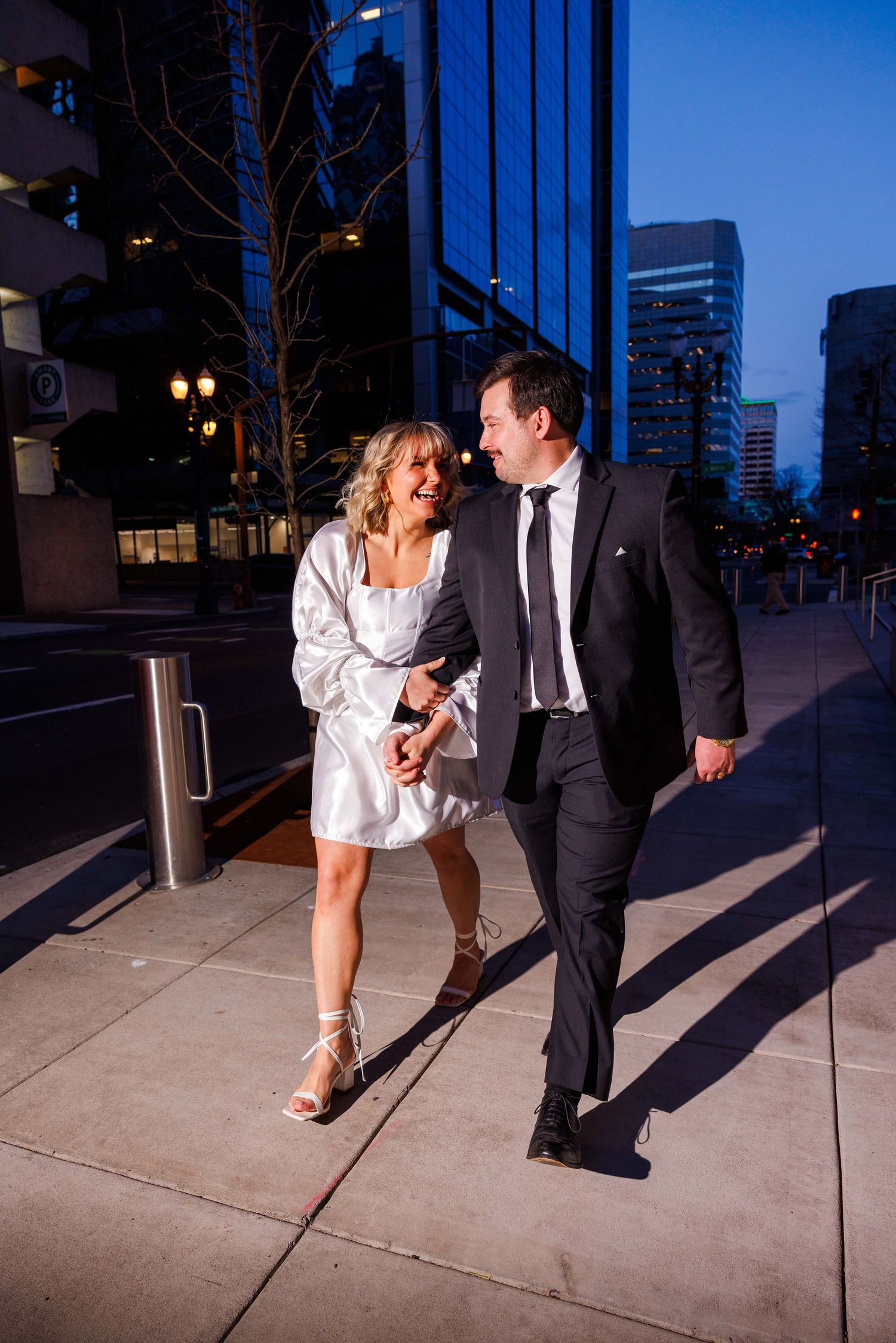 A couple enjoys a romantic evening walk along a city street with illuminated buildings in the background.