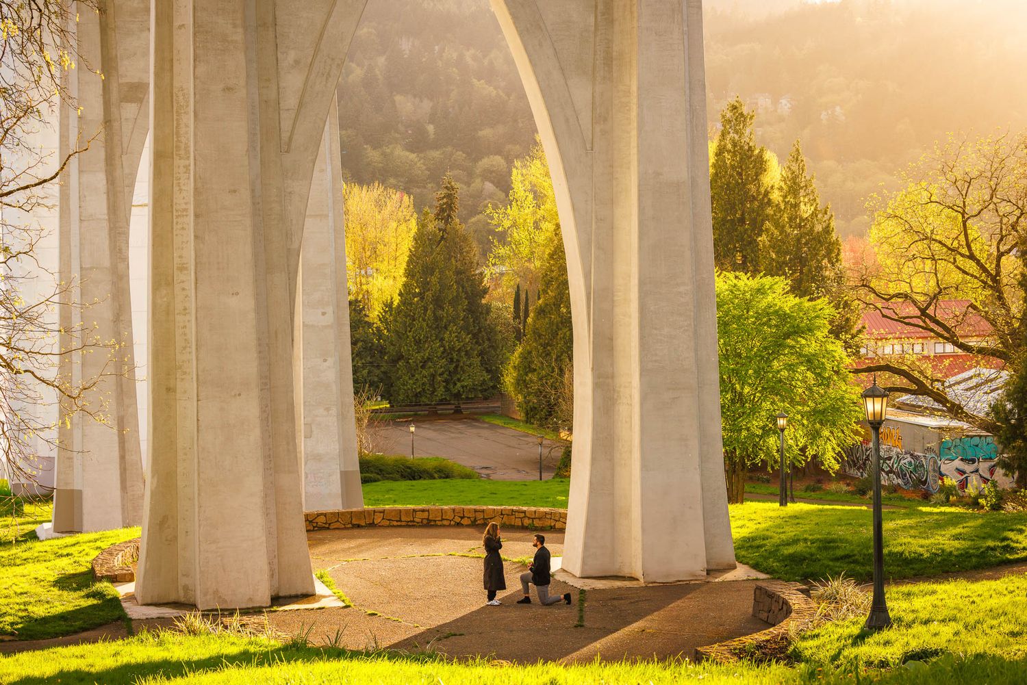 A scenic walkway under massive concrete bridge pillars during golden hour lighting creates dramatic shadows.