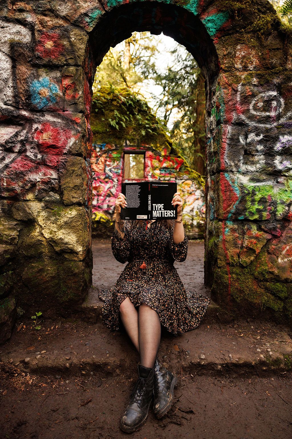 Person in floral dress and boots sits in stone archway covered with colorful graffiti in urban setting.