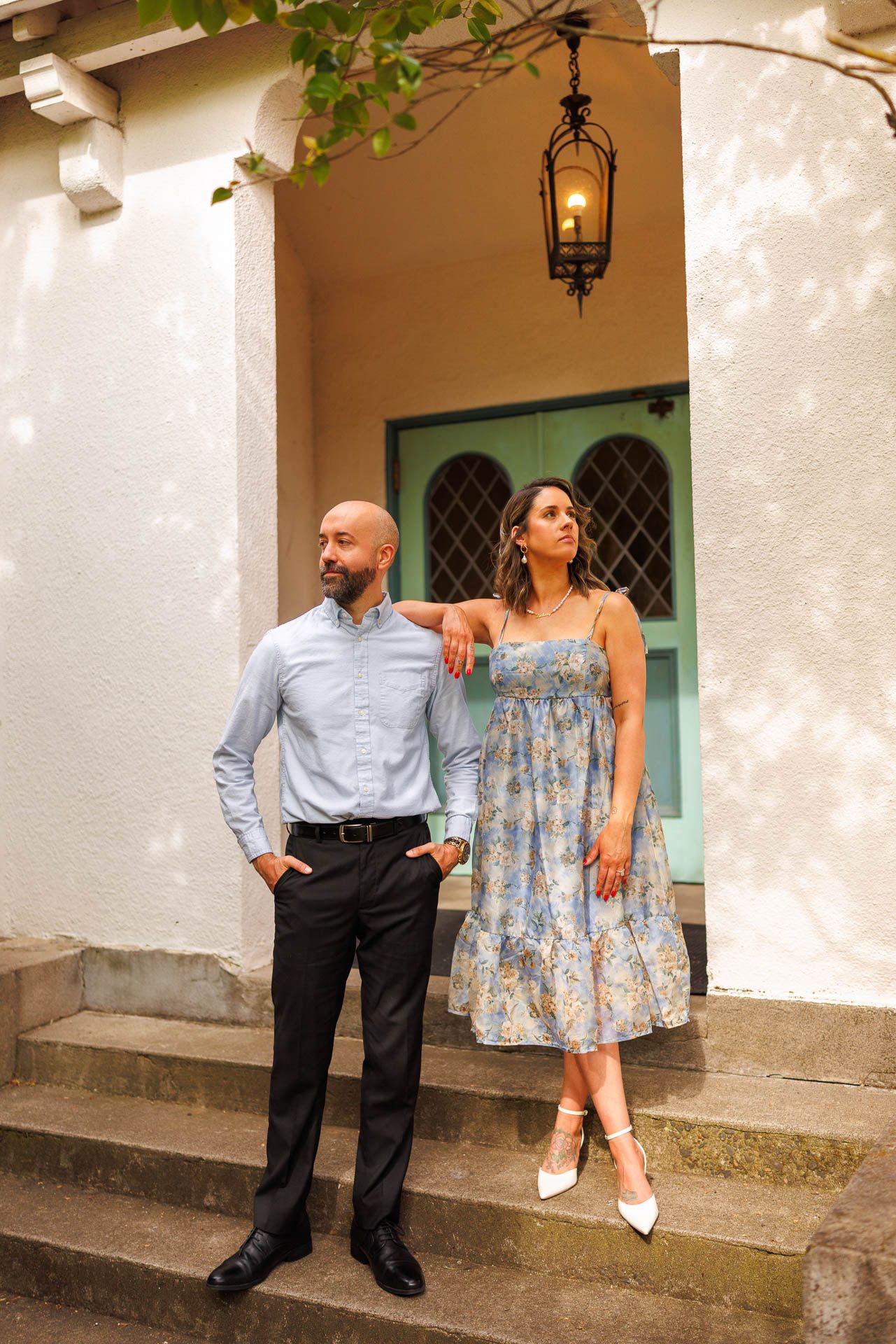 A couple poses on stone steps in front of a teal door, with her in a floral dress and him in business casual attire.