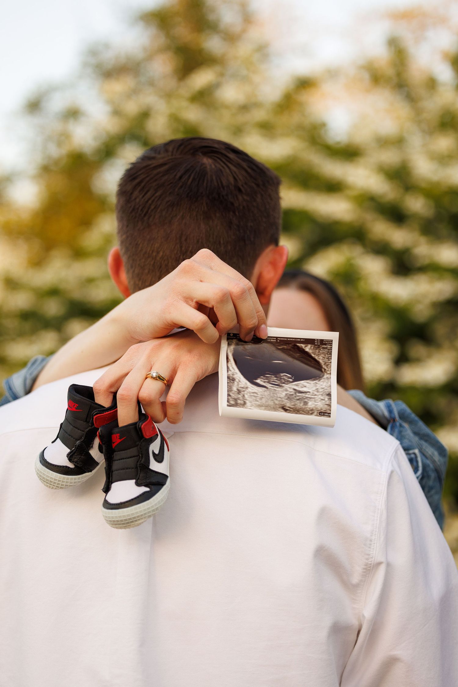 Person holding tiny baby shoes and ultrasound photo against white shirt with trees in soft bokeh background.