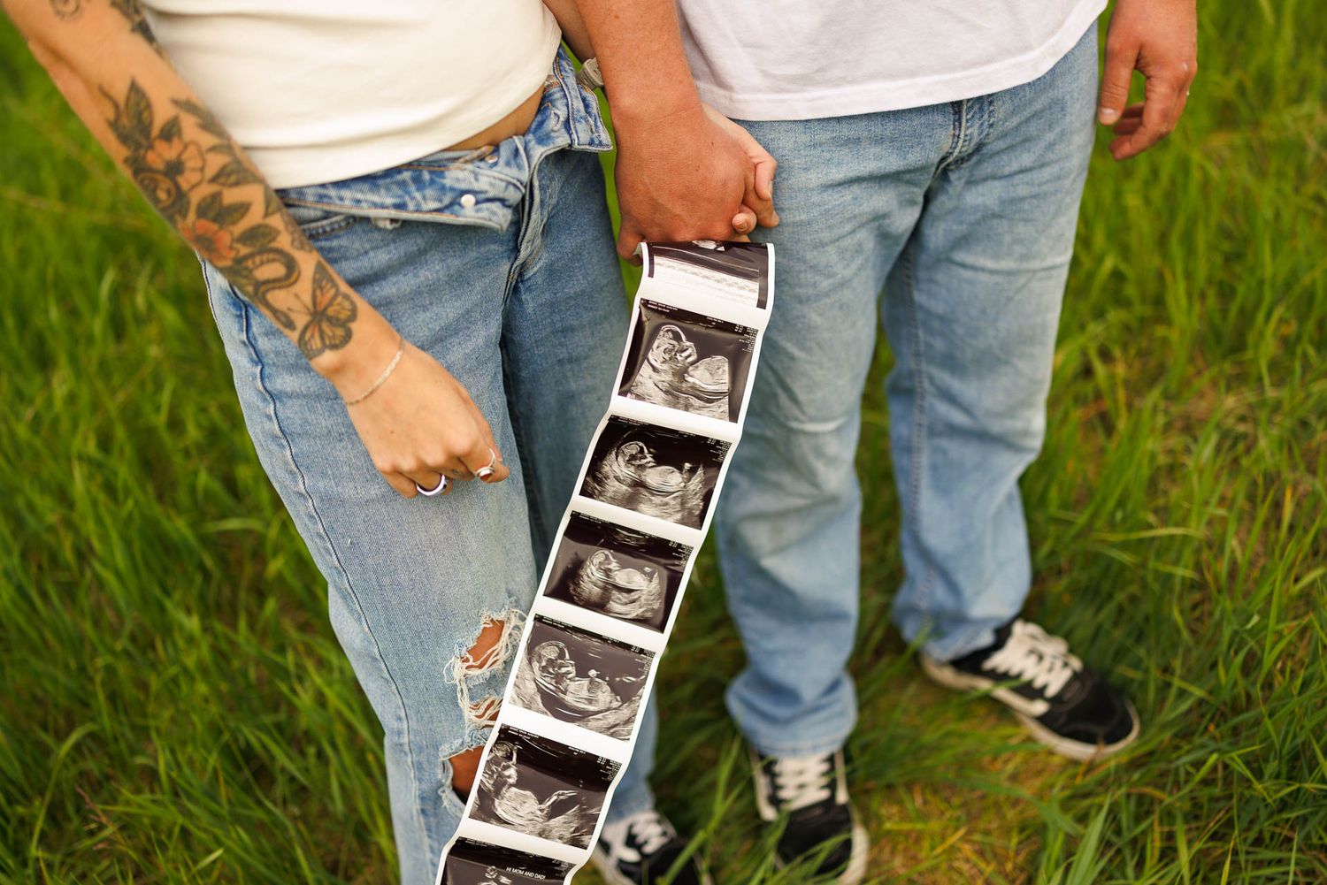 Couple in jeans stands in grass field holding strip of ultrasound images showing their future baby.