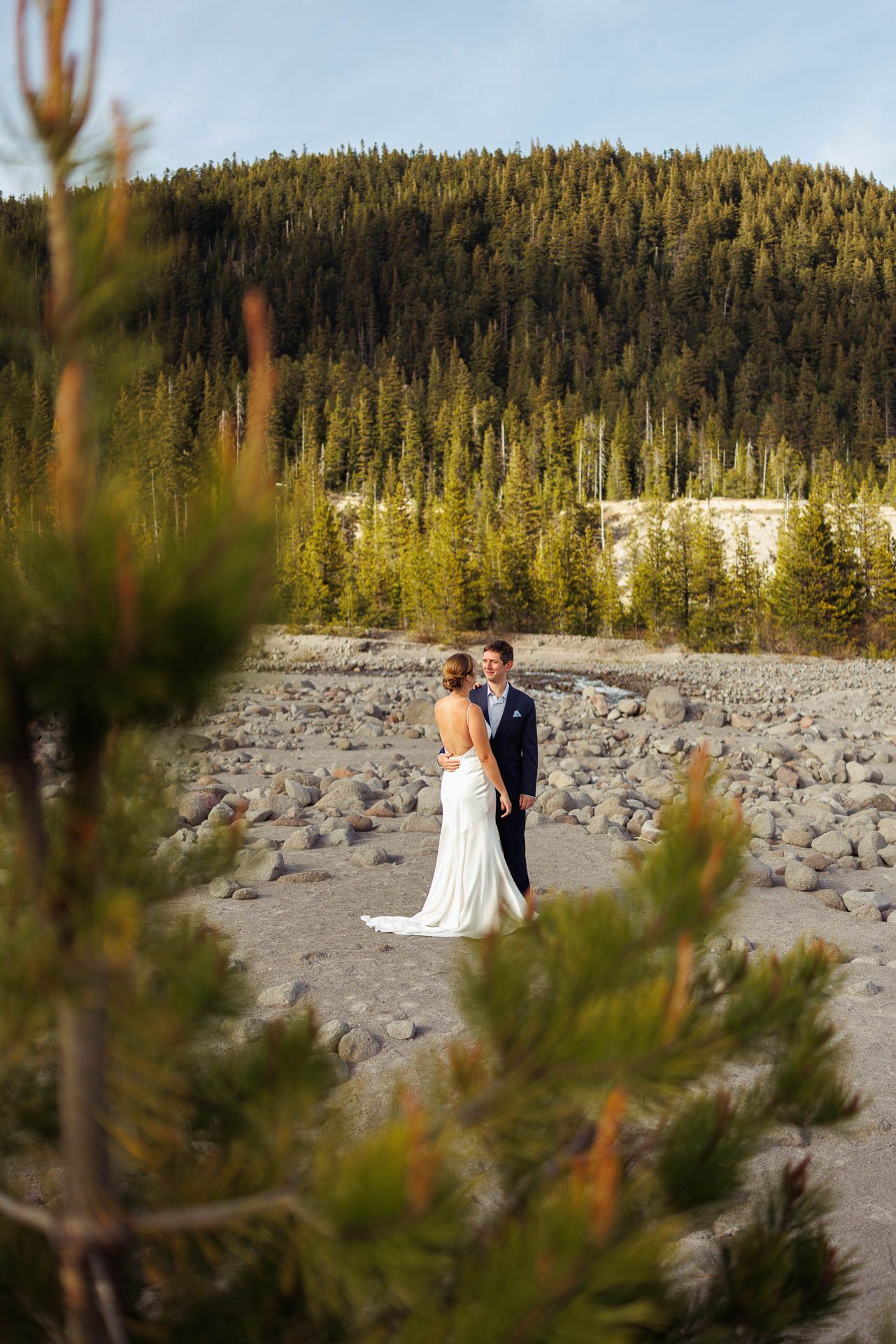 A couple embraces on rocky terrain with evergreen forest-covered mountains in the background during golden hour.