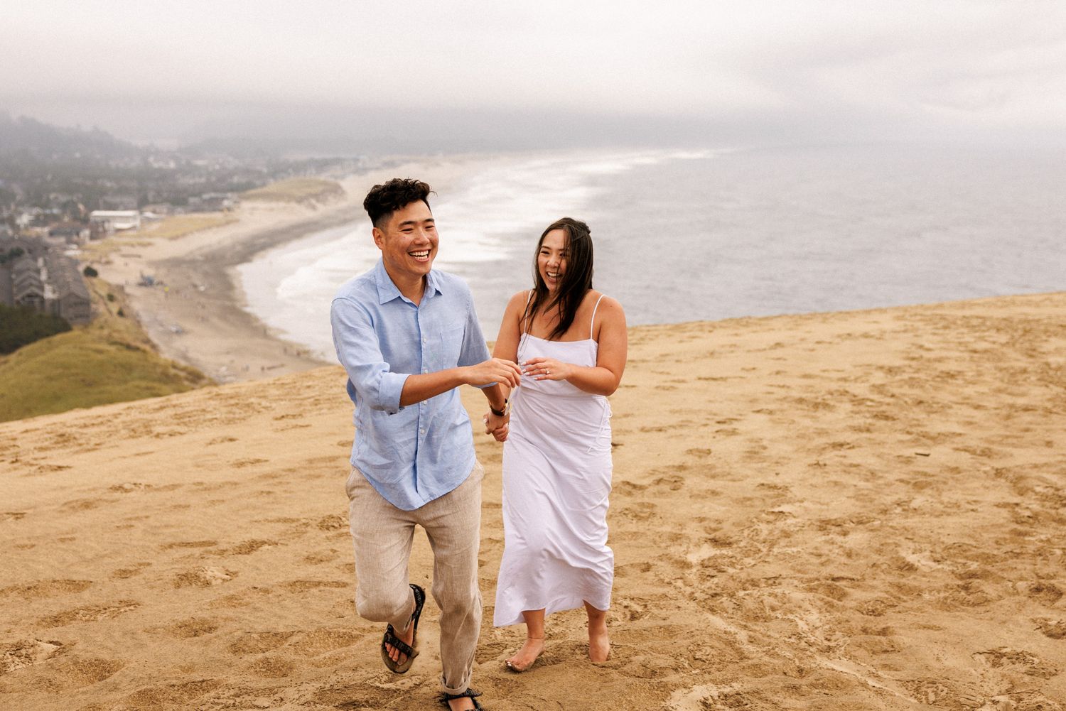 A couple runs together on a sandy hillside overlooking a misty coastal beach on a cloudy day.