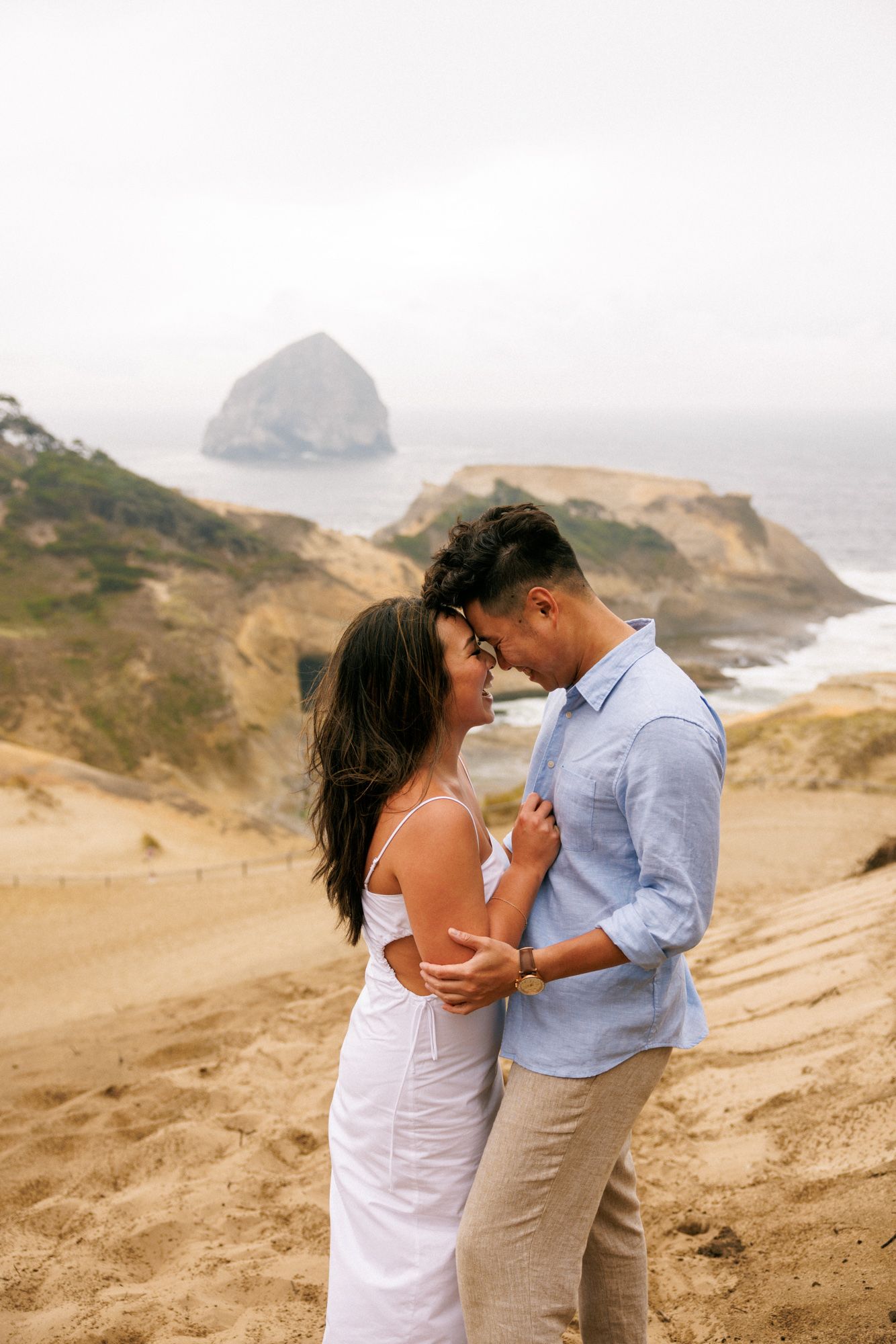 A romantic couple embraces on a cliff overlooking Haystack Rock at Cape Kiwanda on the Oregon Coast during golden hour.