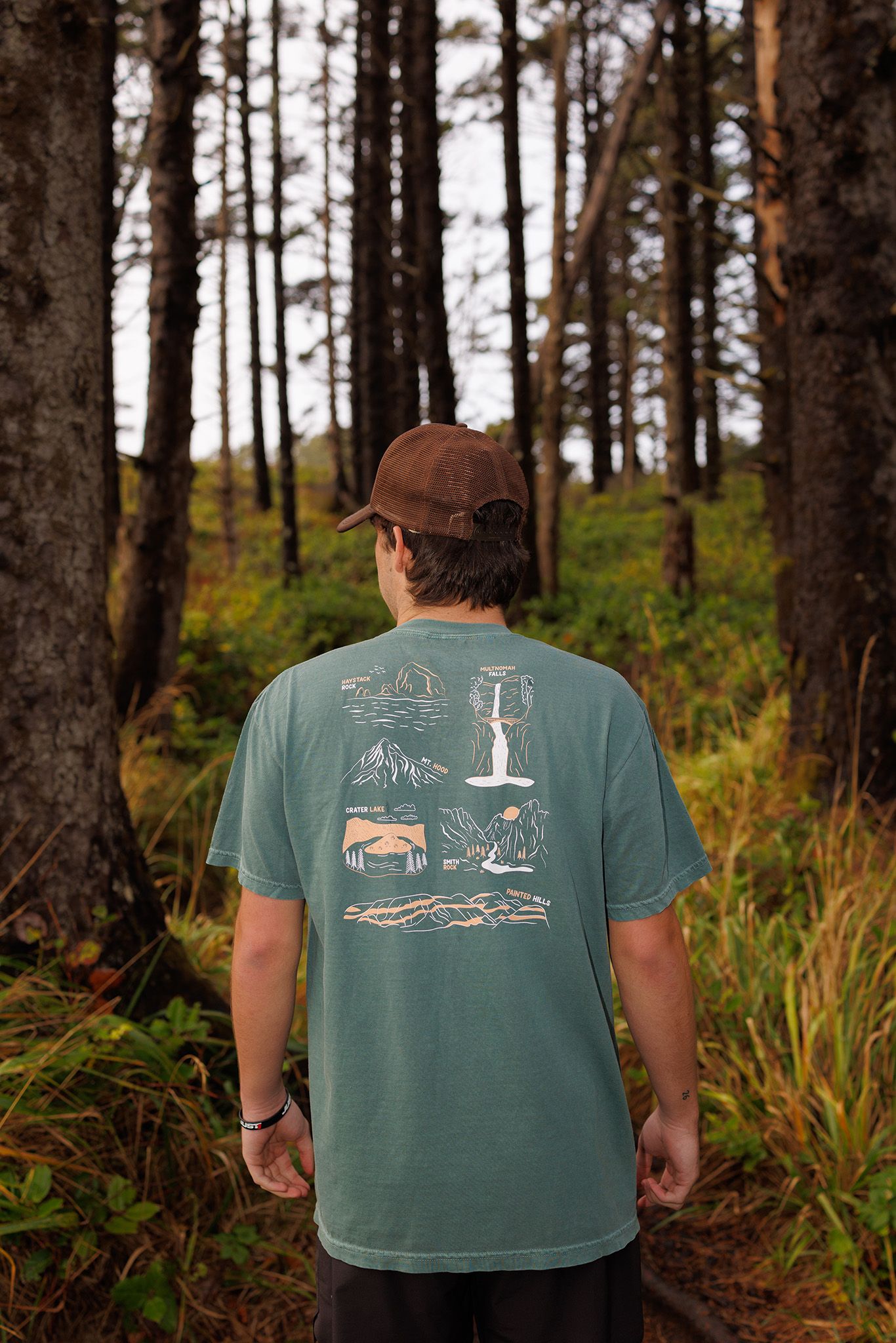 Someone wearing a baseball cap and green t-shirt with outdoor camping artwork stands among tall pine trees in a forest.