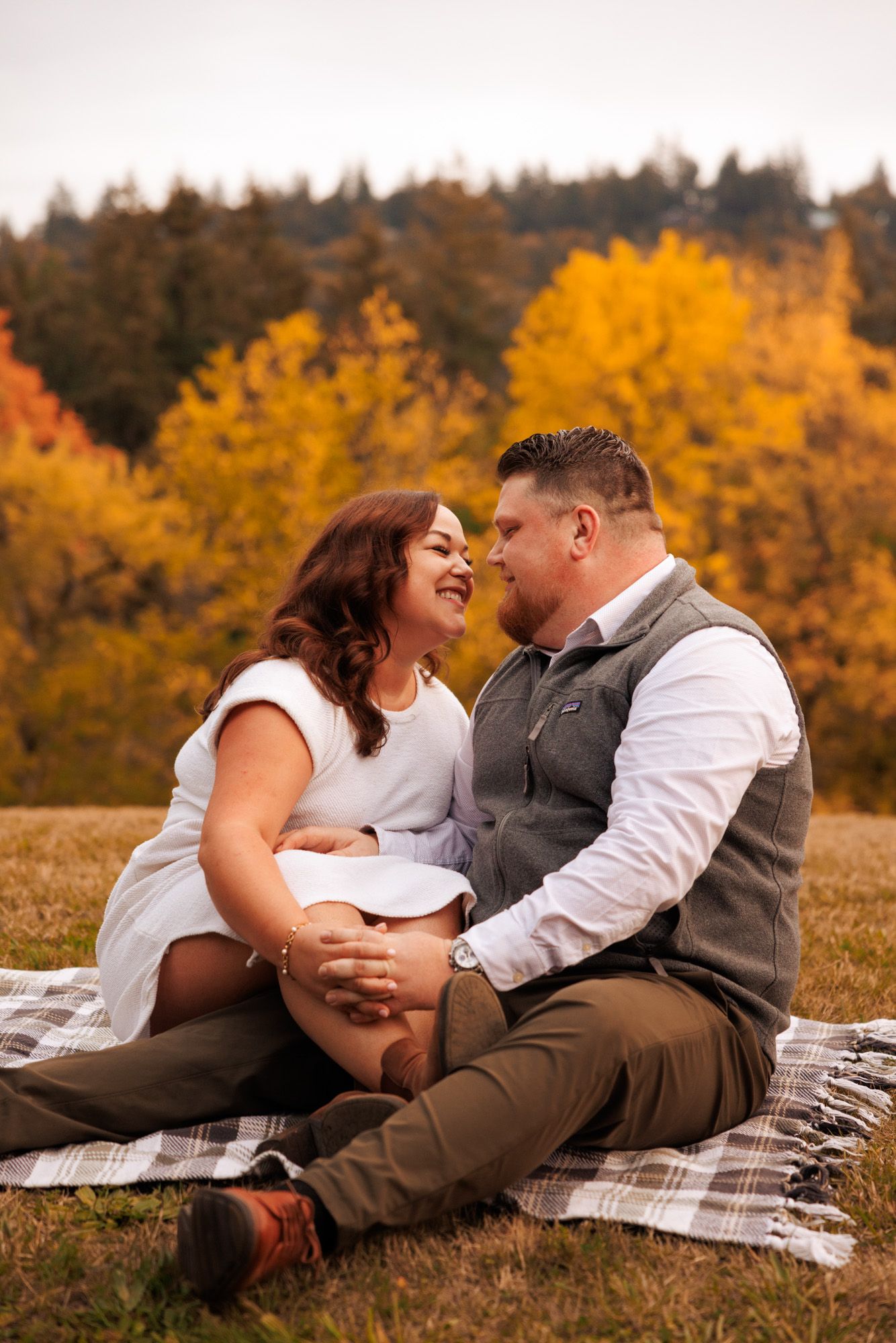 A couple shares a joyful moment on a plaid blanket against vibrant autumn foliage in a mountainside meadow.