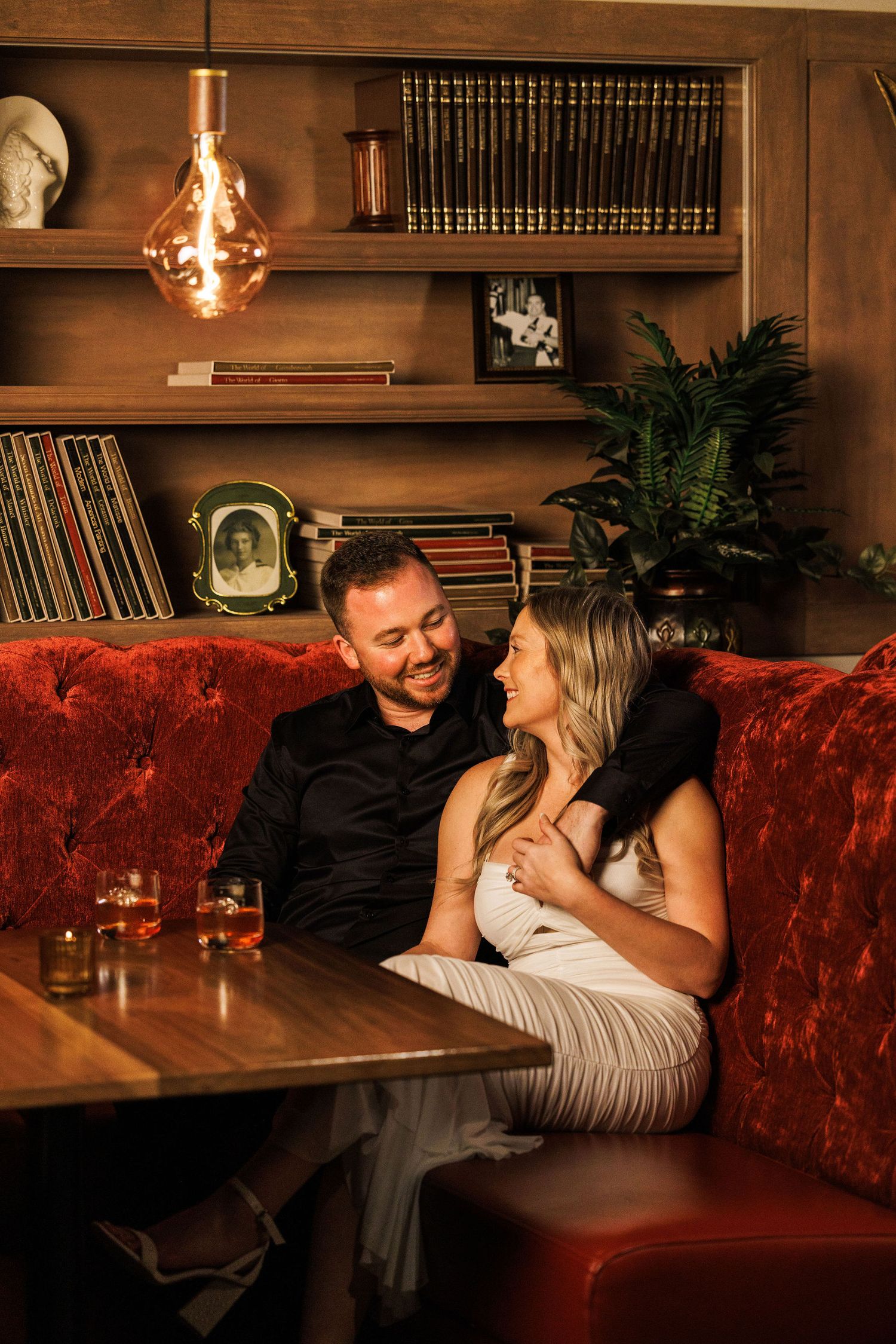 A couple shares an intimate moment on a red velvet couch in a cozy library setting with ambient lighting.