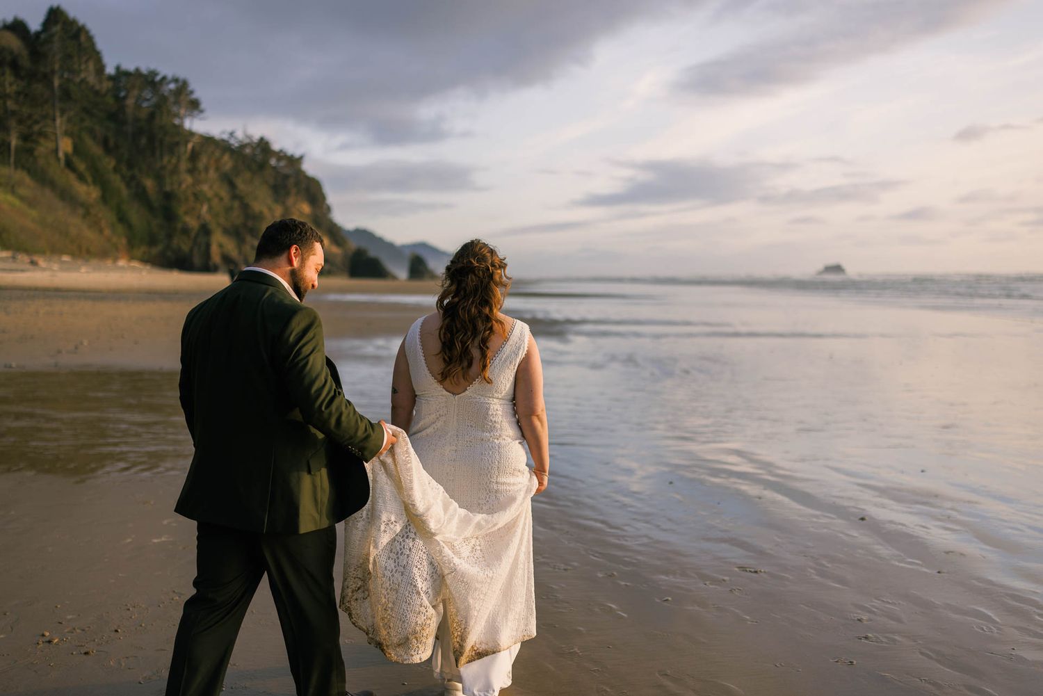 Couple walking along scenic coastal beach at sunset with waves and mountains in background.