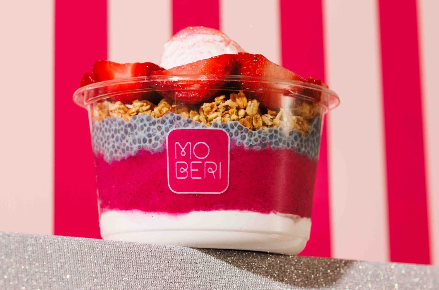 A layered parfait in a clear cup with chia pudding, strawberries, and granola against a pink and white striped background.