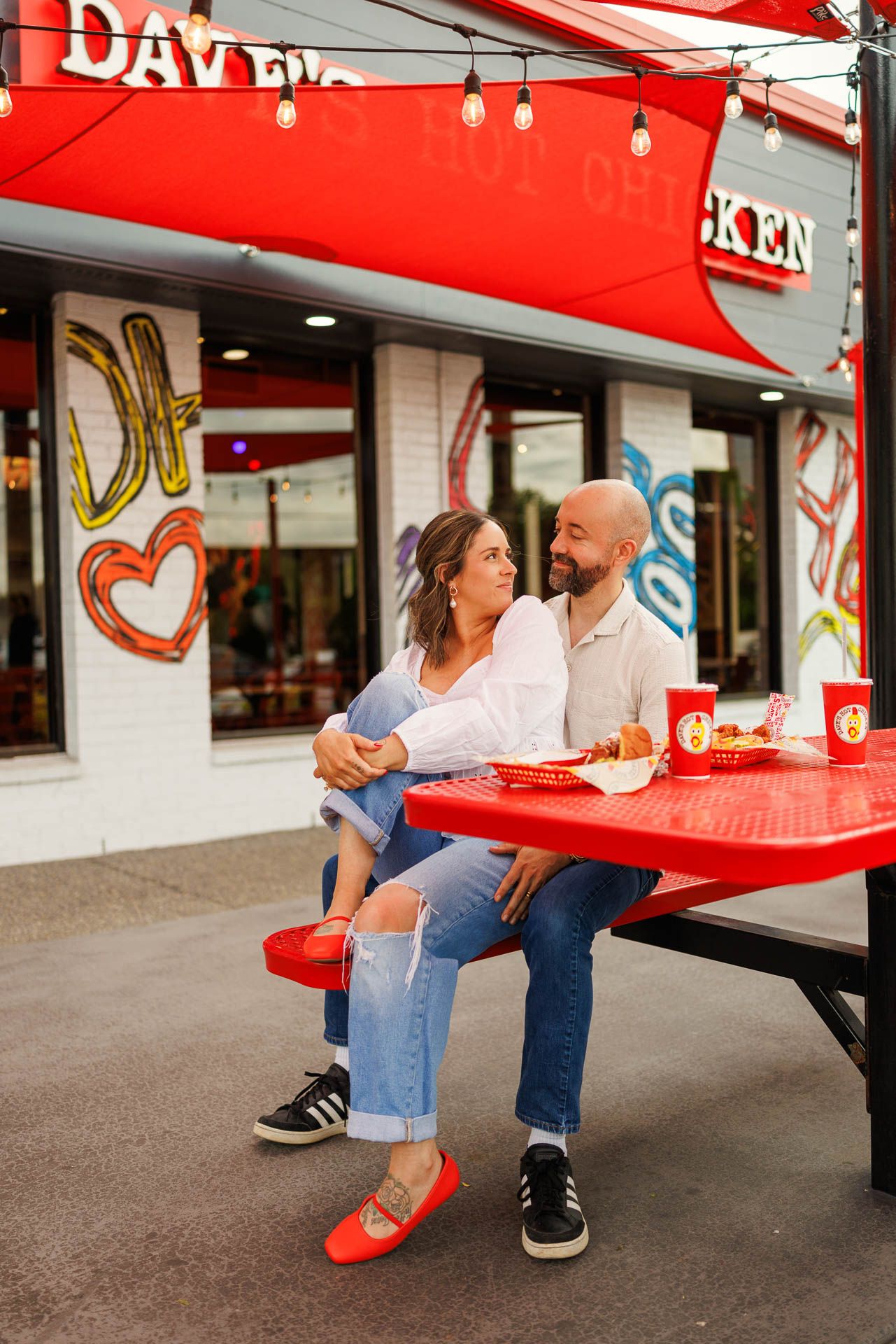 Young couple sitting at red retro diner tables enjoy a casual meal together under string lights.