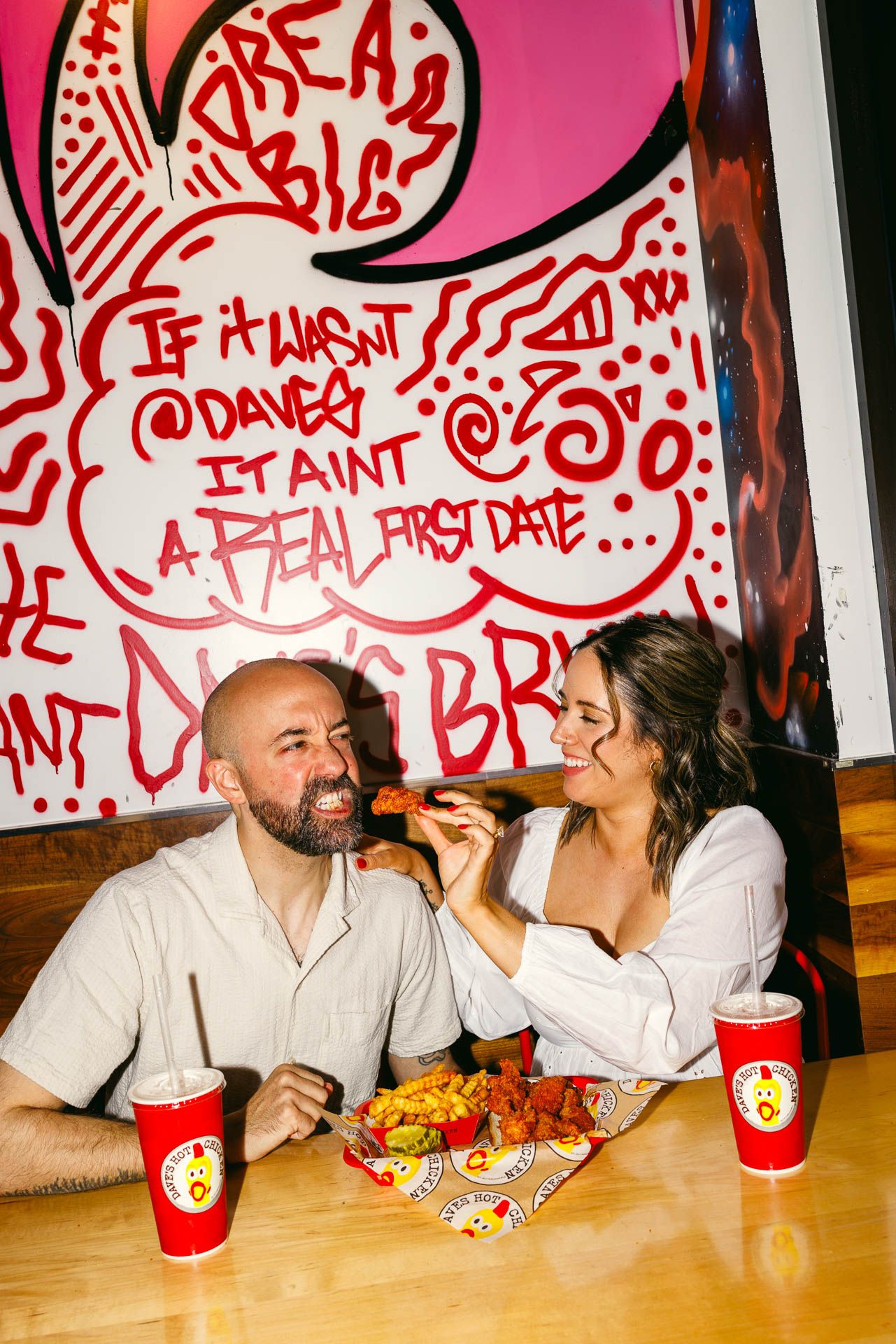 Two people sharing food and enjoying drinks with red cups at a restaurant table with graffiti art on the wall behind them.