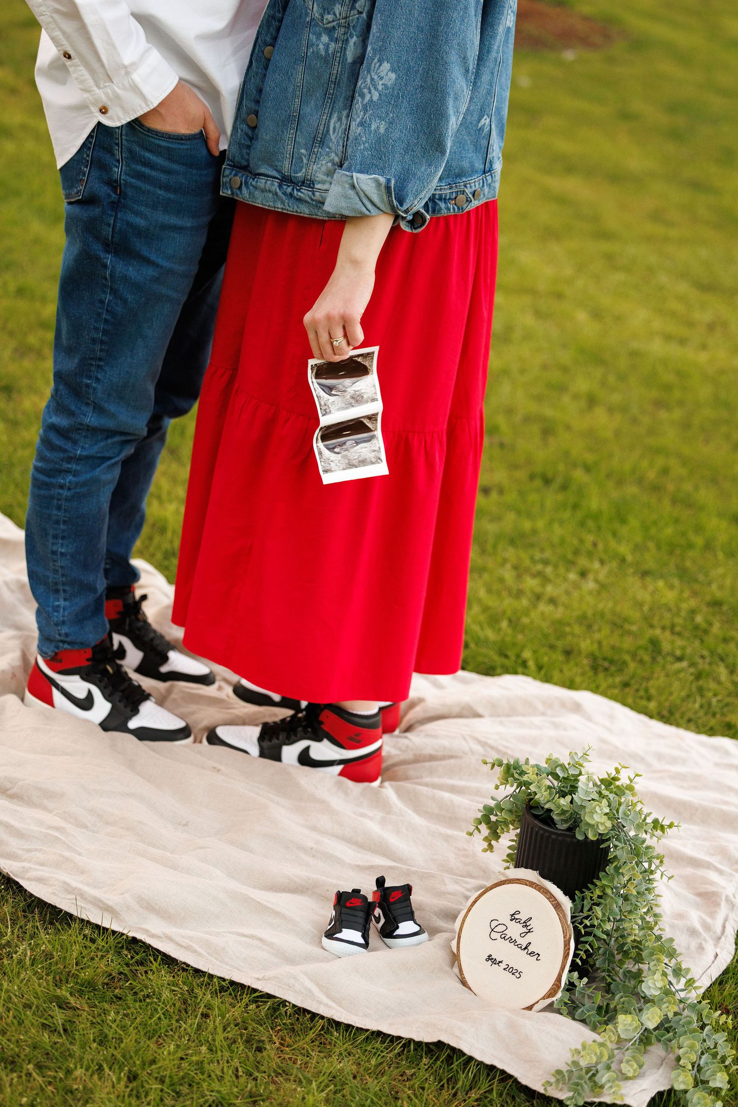 Couple stands on blanket with baby shoes and foliage, wearing denim, red skirt and matching Nike sneakers.