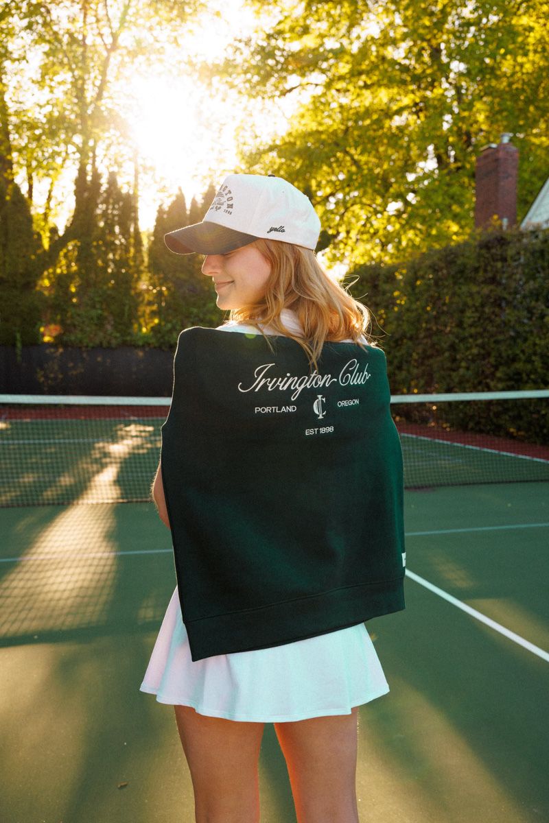 A tennis player stands on a green court at sunset with long shadows and golden sunlight filtering through the trees.