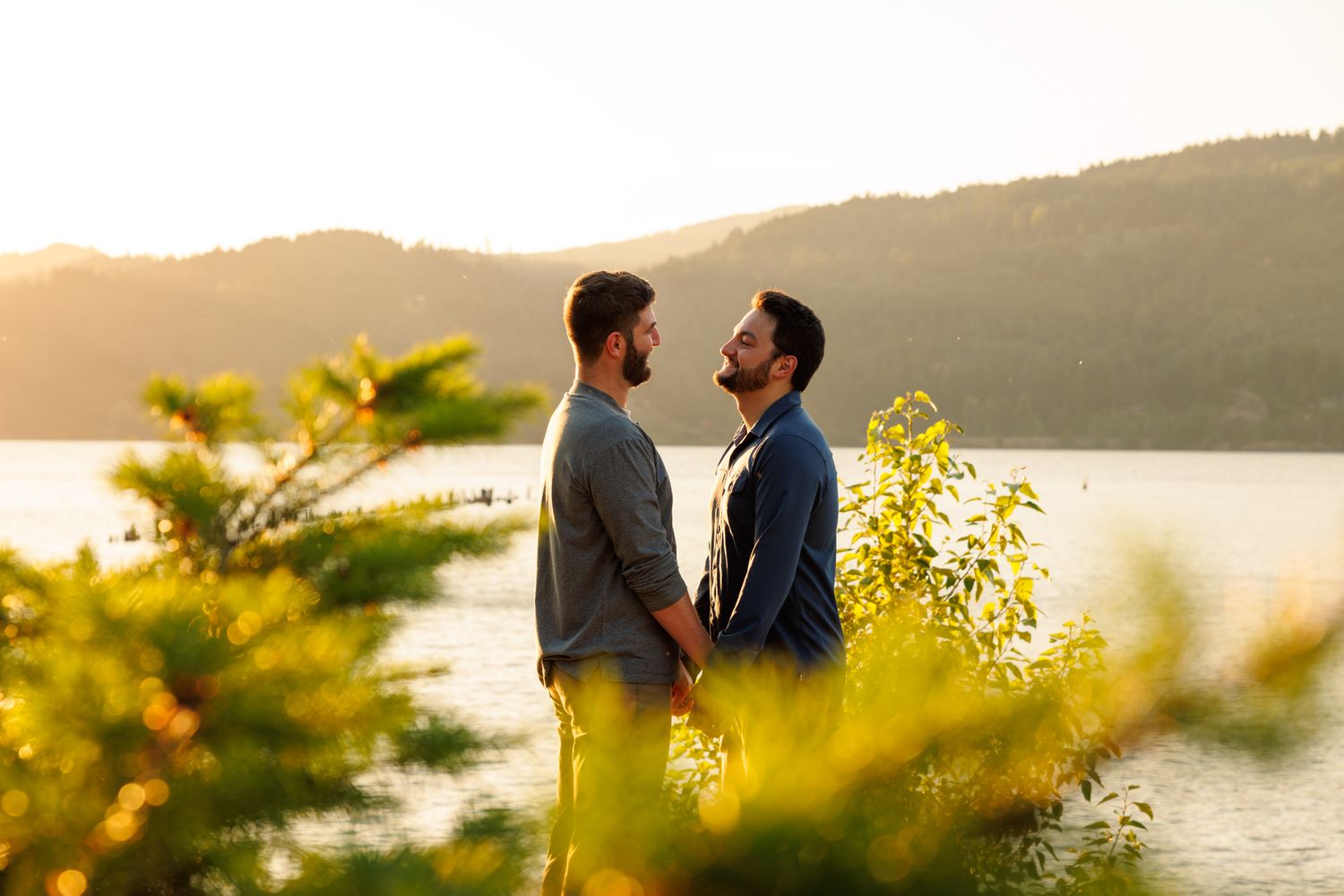 A couple shares a romantic moment at sunset by a lake, surrounded by golden-lit foliage and mountain scenery.