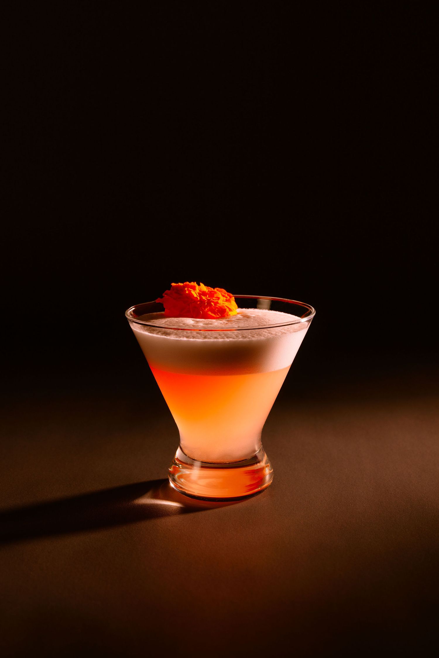 Elegant layered cocktail with orange garnish in martini glass against dark background with dramatic lighting.