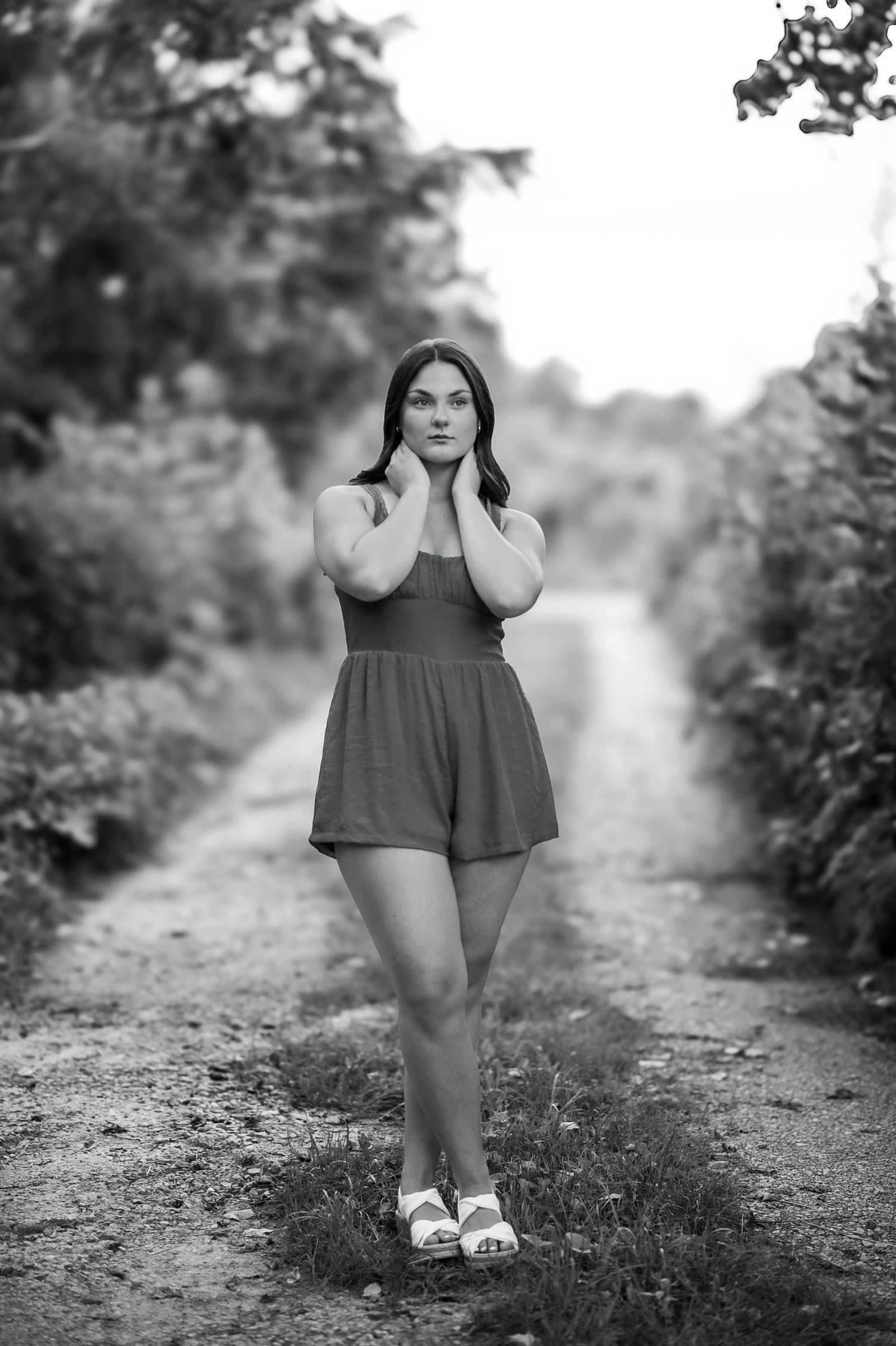 Black and white portrait photograph of a person in a romper standing on a rural dirt path surrounded by greenery.
