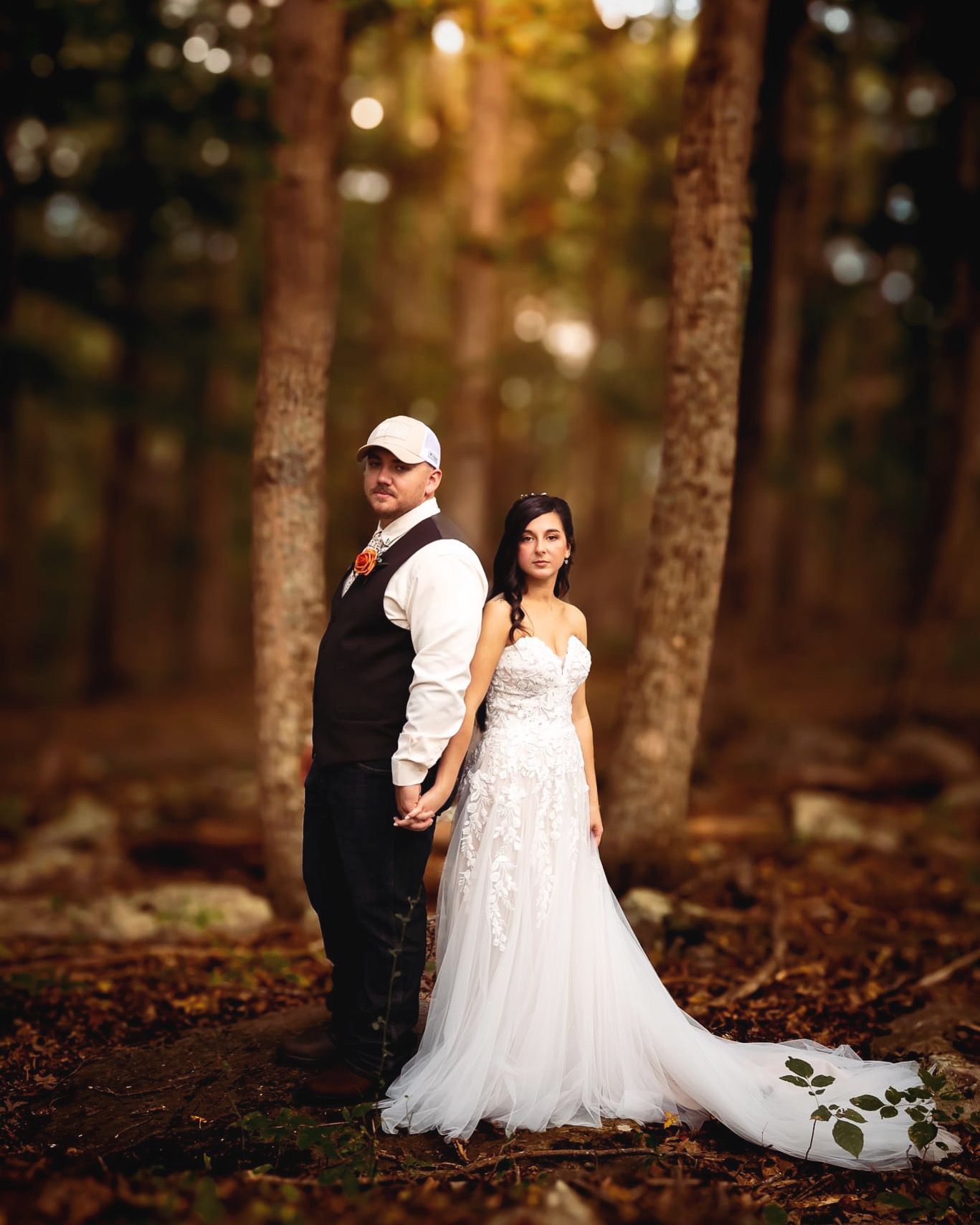 A rustic woodland wedding photo with a couple posing between tall trees in natural lighting.