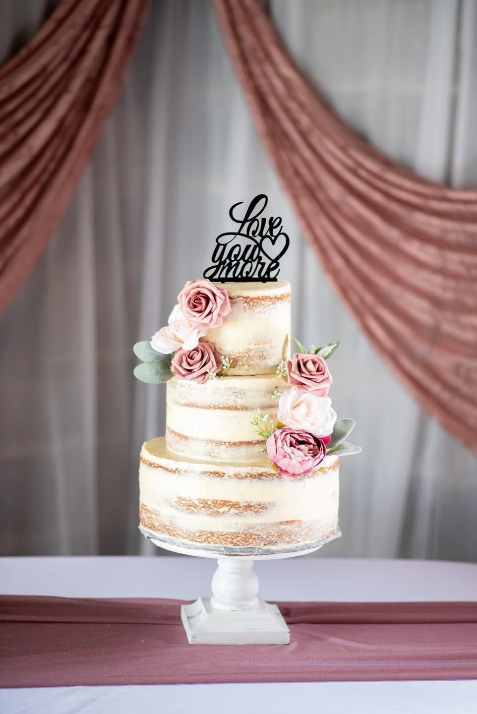 Elegant semi-naked wedding cake decorated with pink roses and 'Love you more' topper.