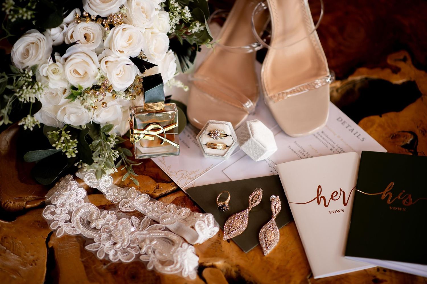 Wedding details including white roses, bridal shoes, and vows books arranged on a wooden surface.
