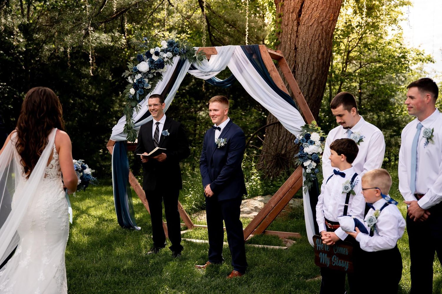 Wedding ceremony under wooden arch with white draping in forest setting.