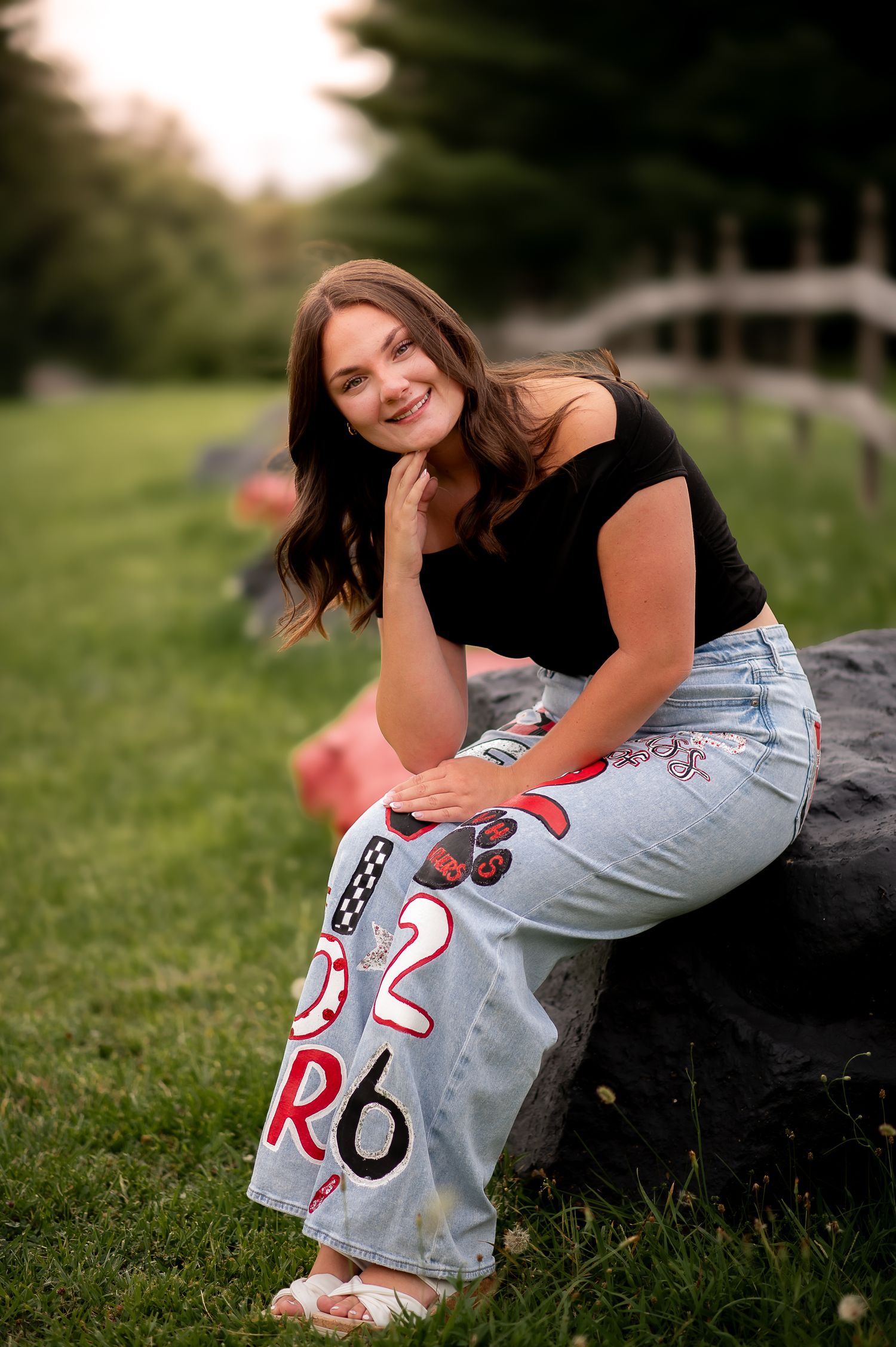 A person wearing a black top and embellished jeans poses casually on a rock with a blurred nature background at sunset.