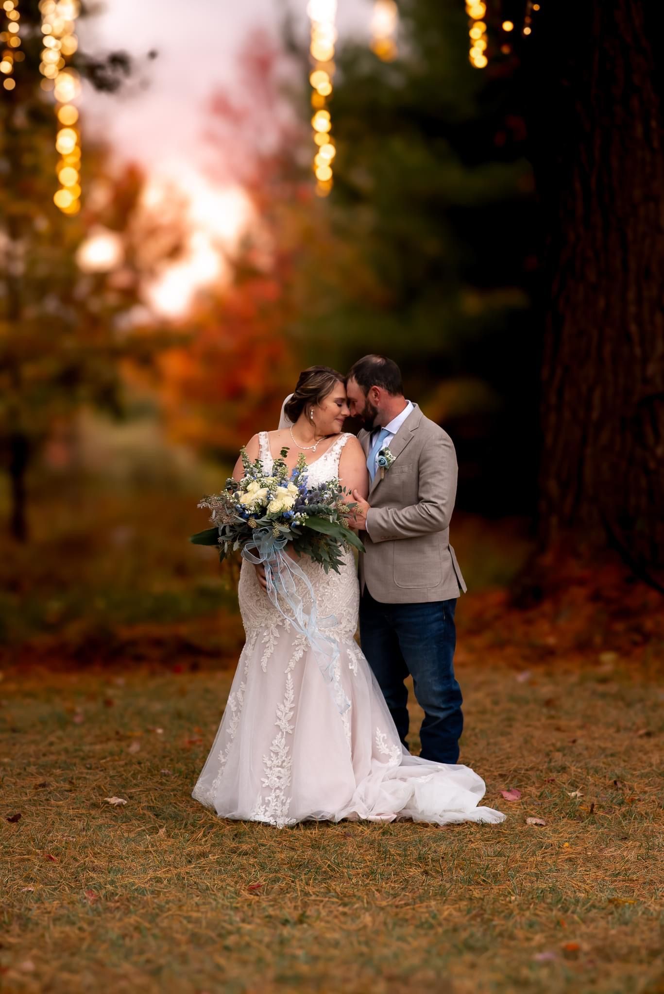 Romantic fall wedding photo session with bokeh lights in wooded setting shows couple embracing on autumn ground.