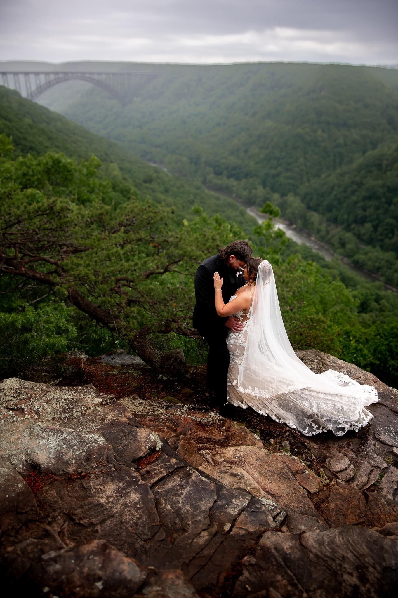 A romantic wedding moment set against a dramatic mountain landscape with a bridge in the distance.
