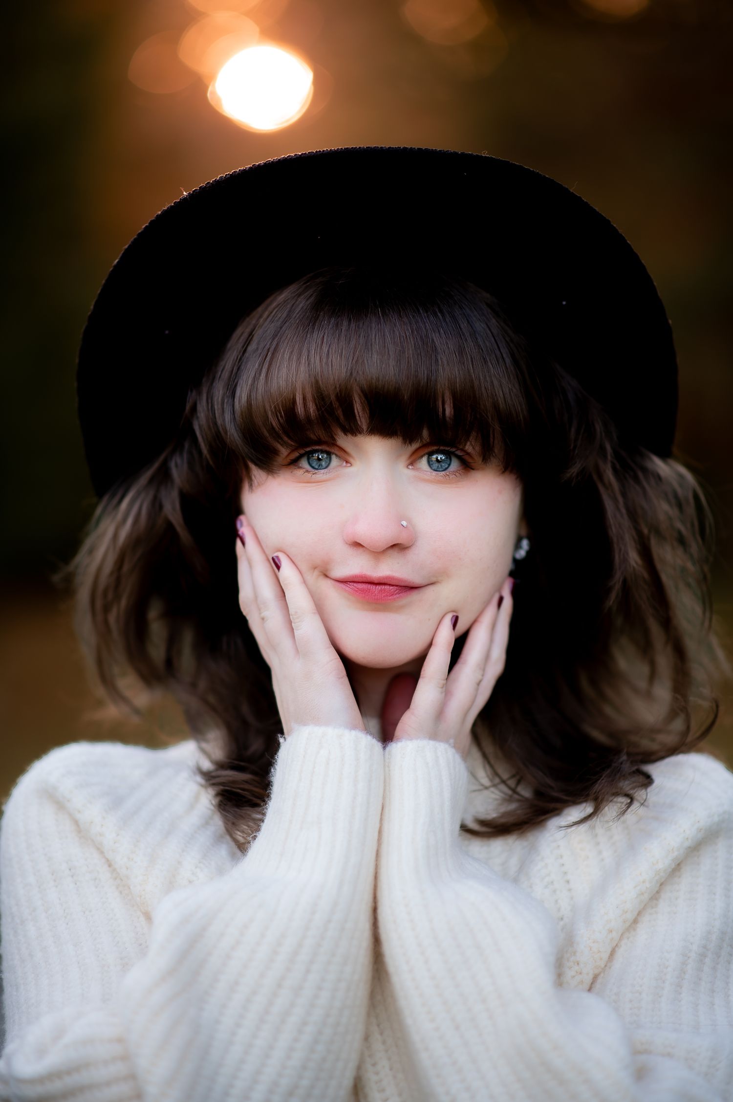 Portrait wearing black wide-brim hat and white sweater with warm bokeh lighting.