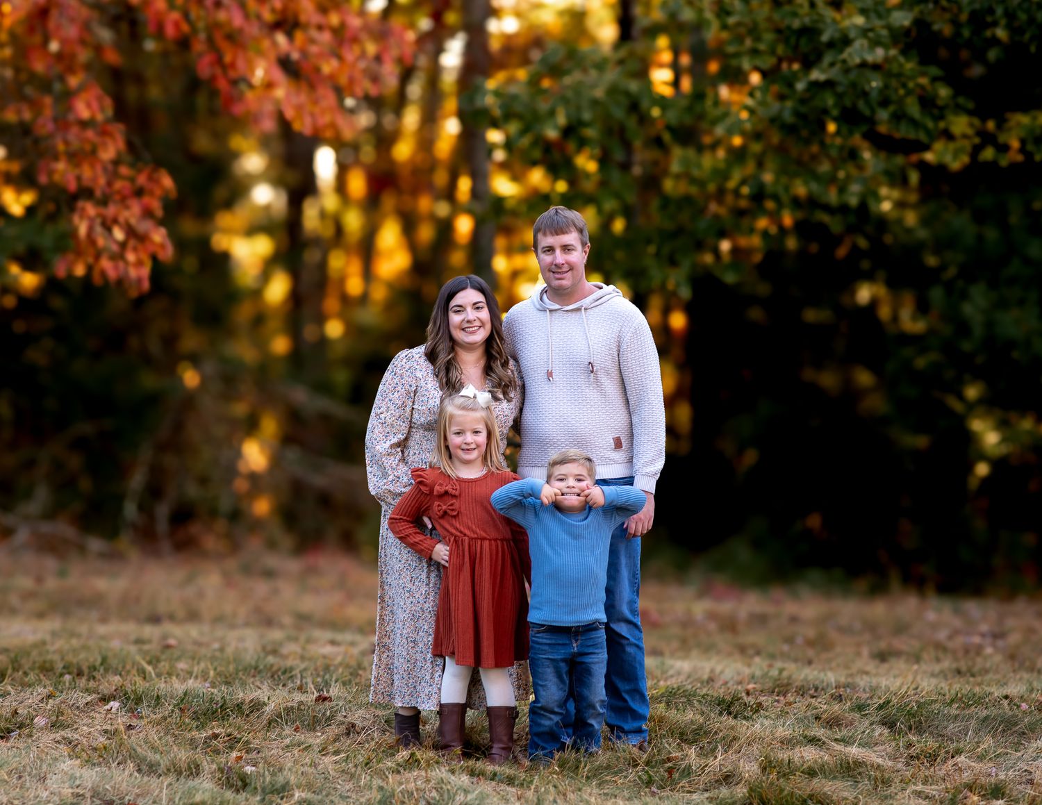 Family of four poses together on grassy hill during autumn sunset.