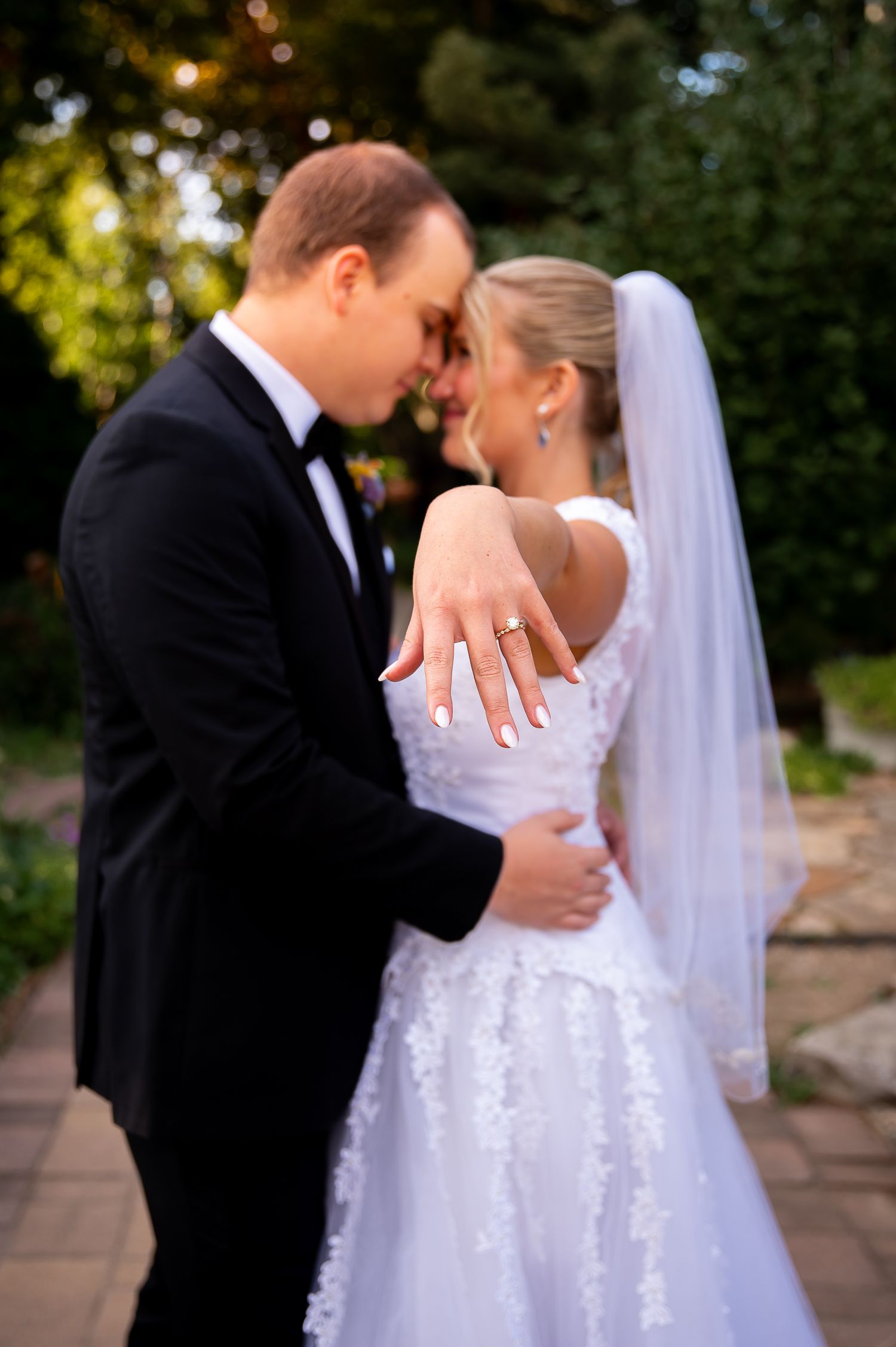 A romantic wedding couple embraces in a garden setting, with the bride wearing a white lace dress and flowing veil.