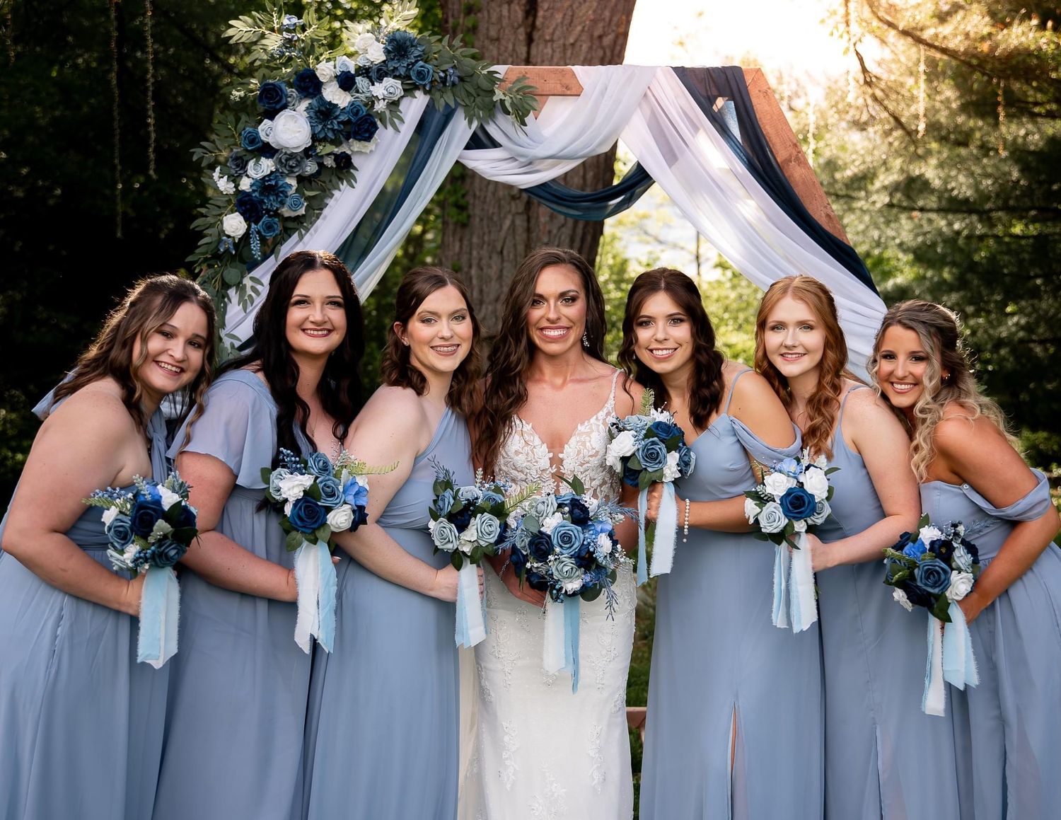 Wedding party in light blue bridesmaid dresses poses under floral arch with navy accents.