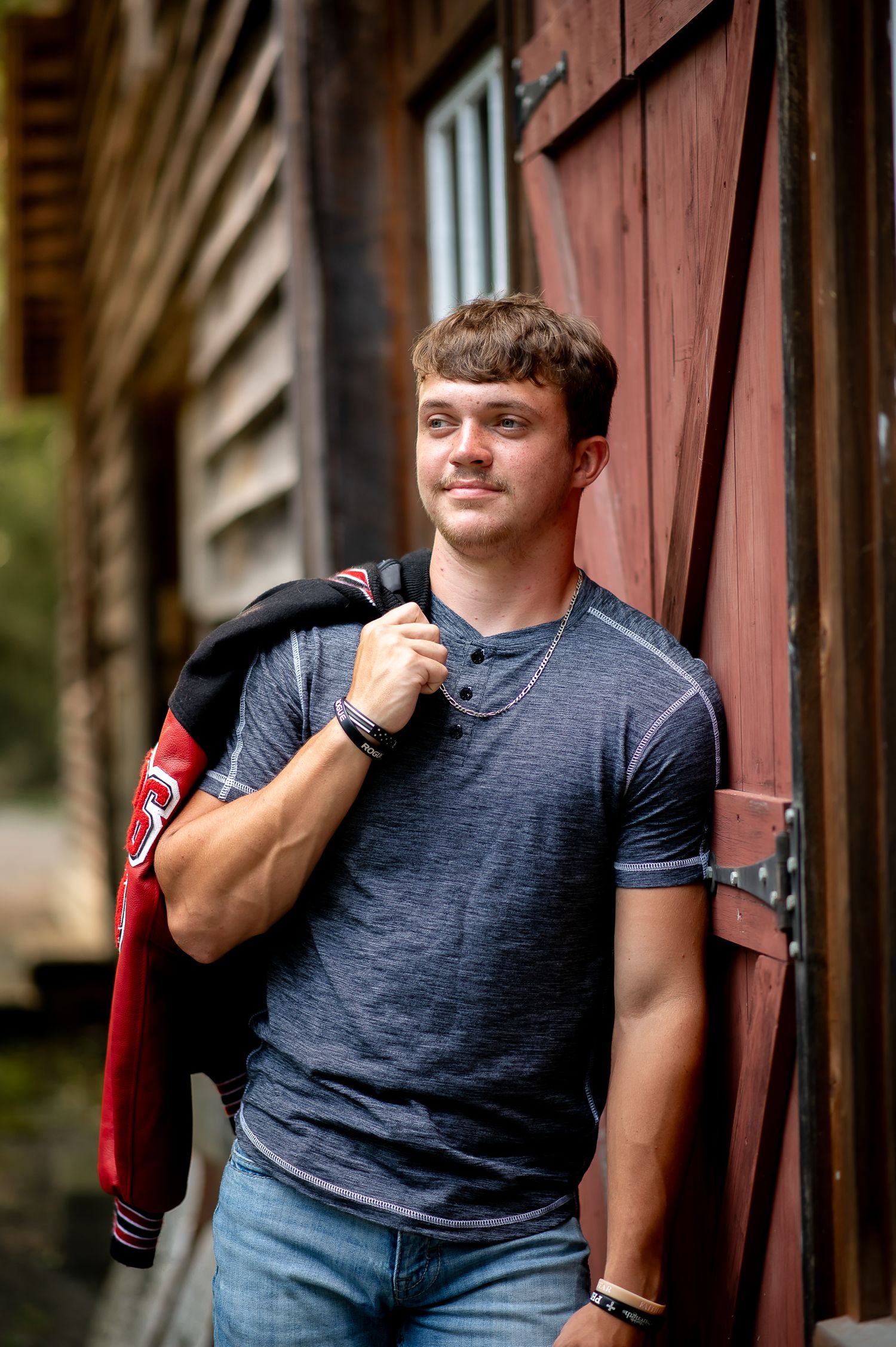 A casual portrait against a rustic red barn door wearing a gray t-shirt and carrying a jacket.
