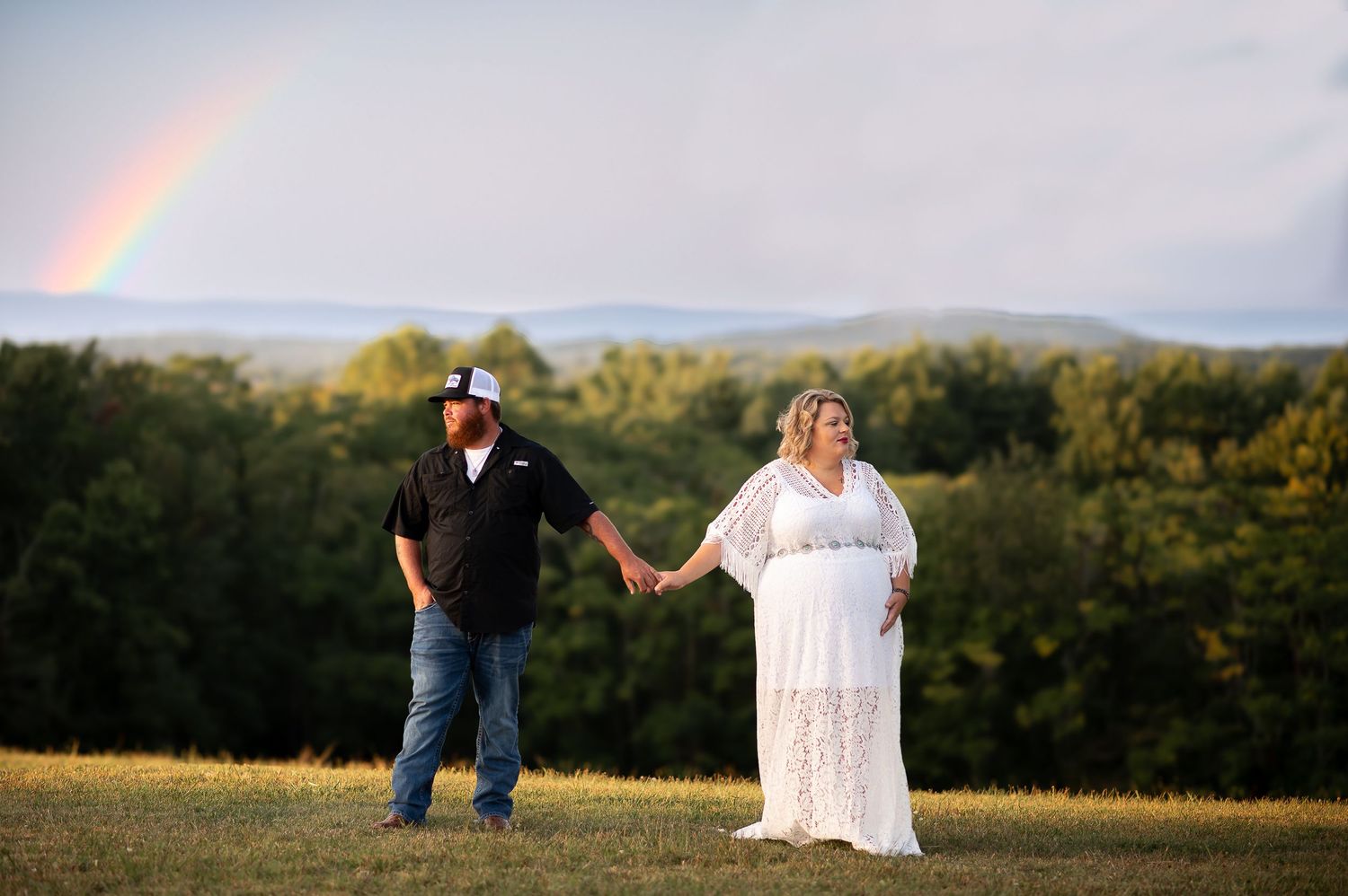 Couple holds hands in scenic hilltop setting with rainbow in background.
