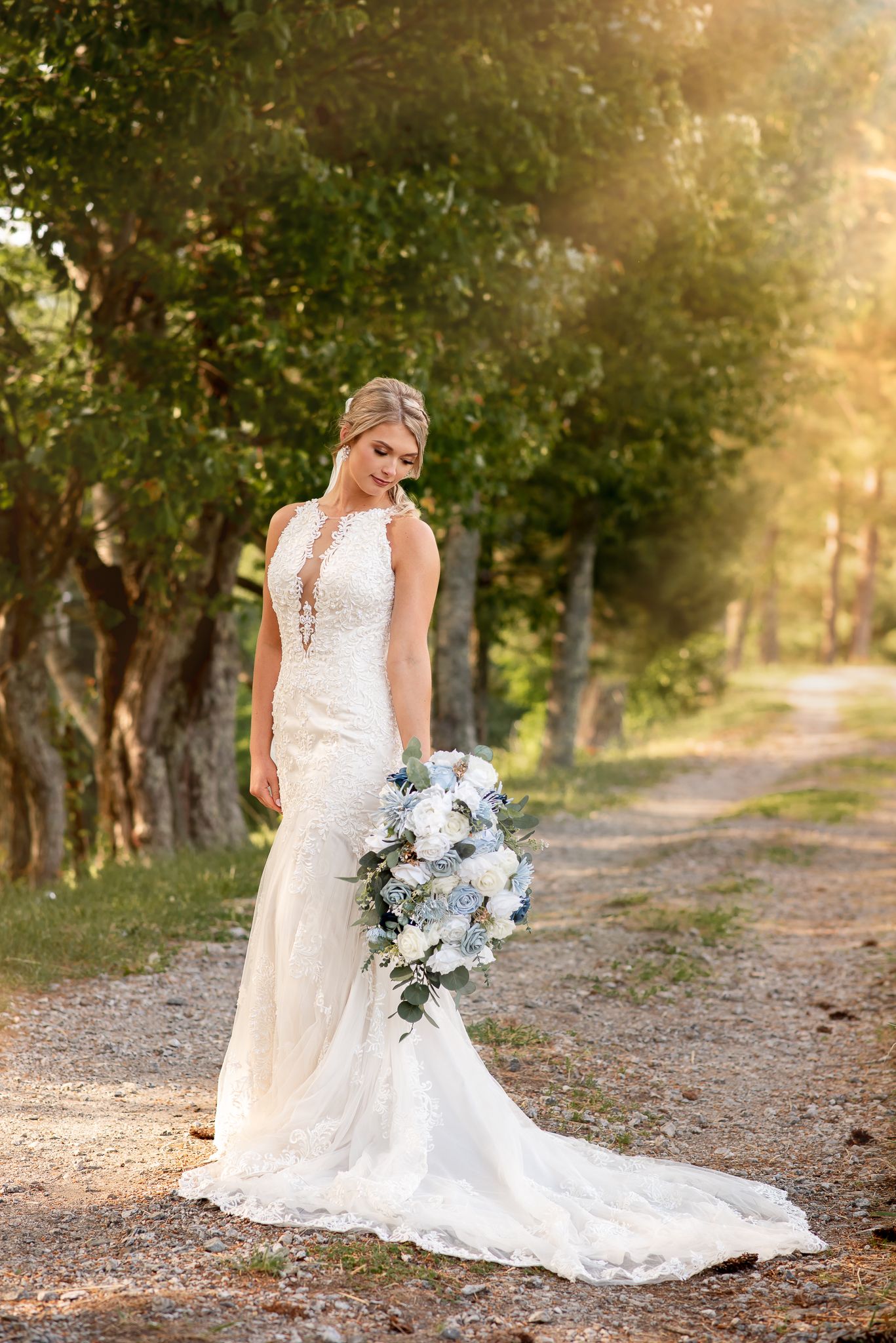 Bride in a fitted lace wedding dress holds a blue and white bouquet on a sunlit forest path.