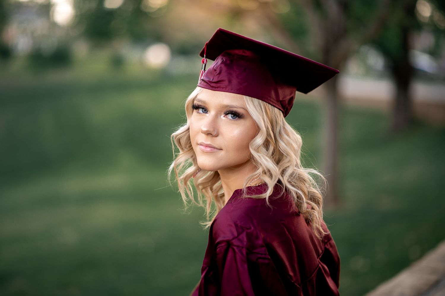 Graduate in burgundy cap and gown poses outdoors with soft bokeh background.