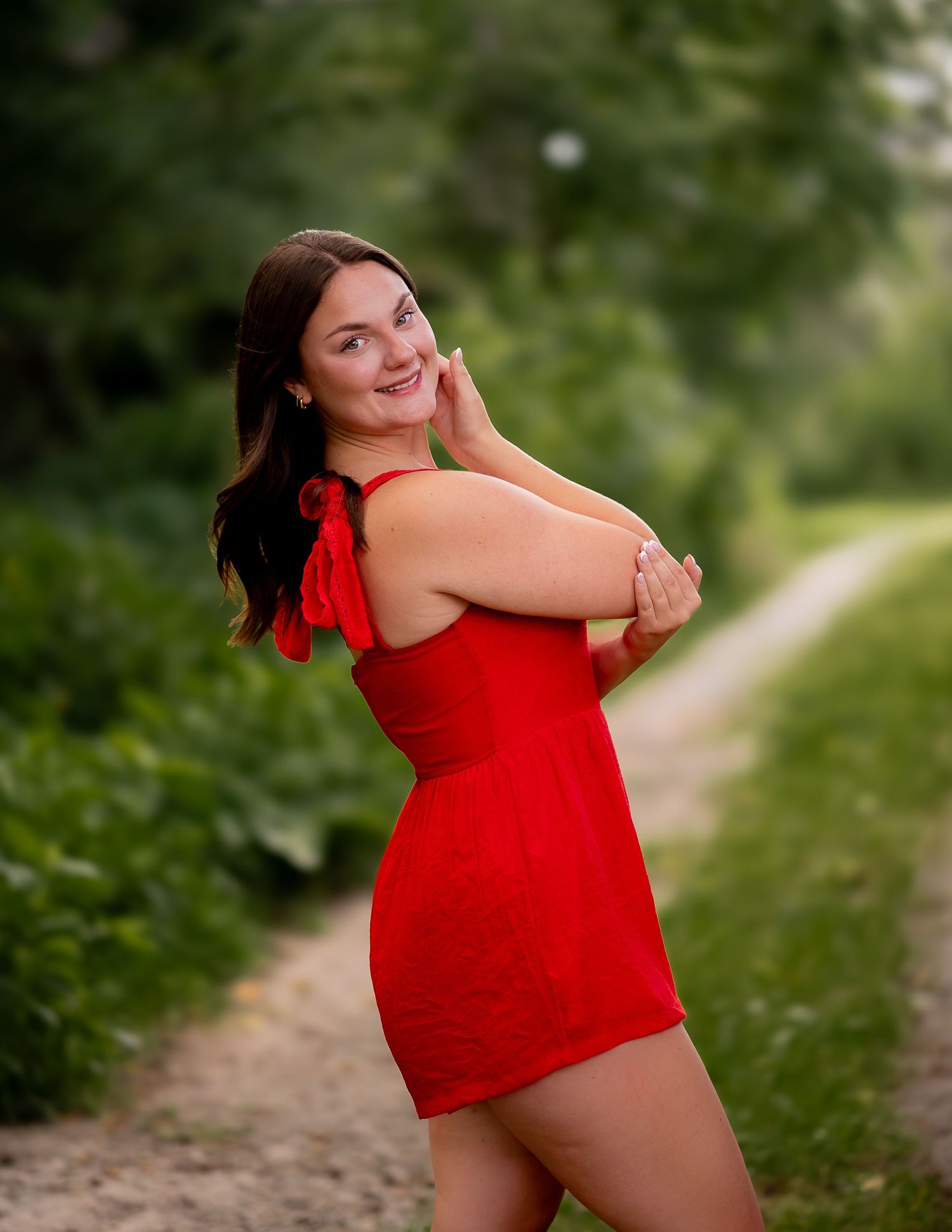 Woman in a vibrant red dress poses cheerfully on a garden path surrounded by lush greenery.
